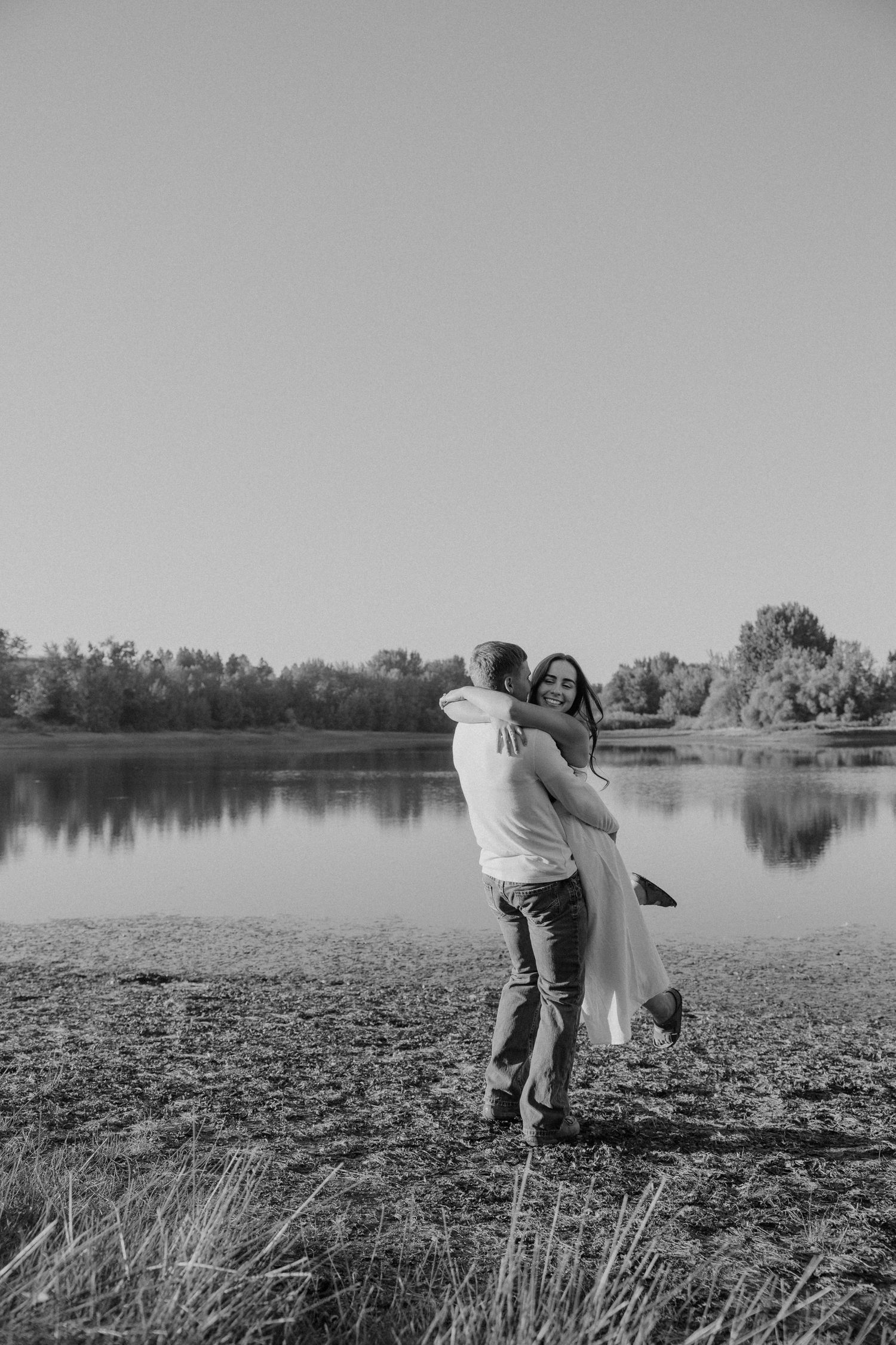 A romantic black and white photo series shows a couple embracing by a serene lake with trees in the background.