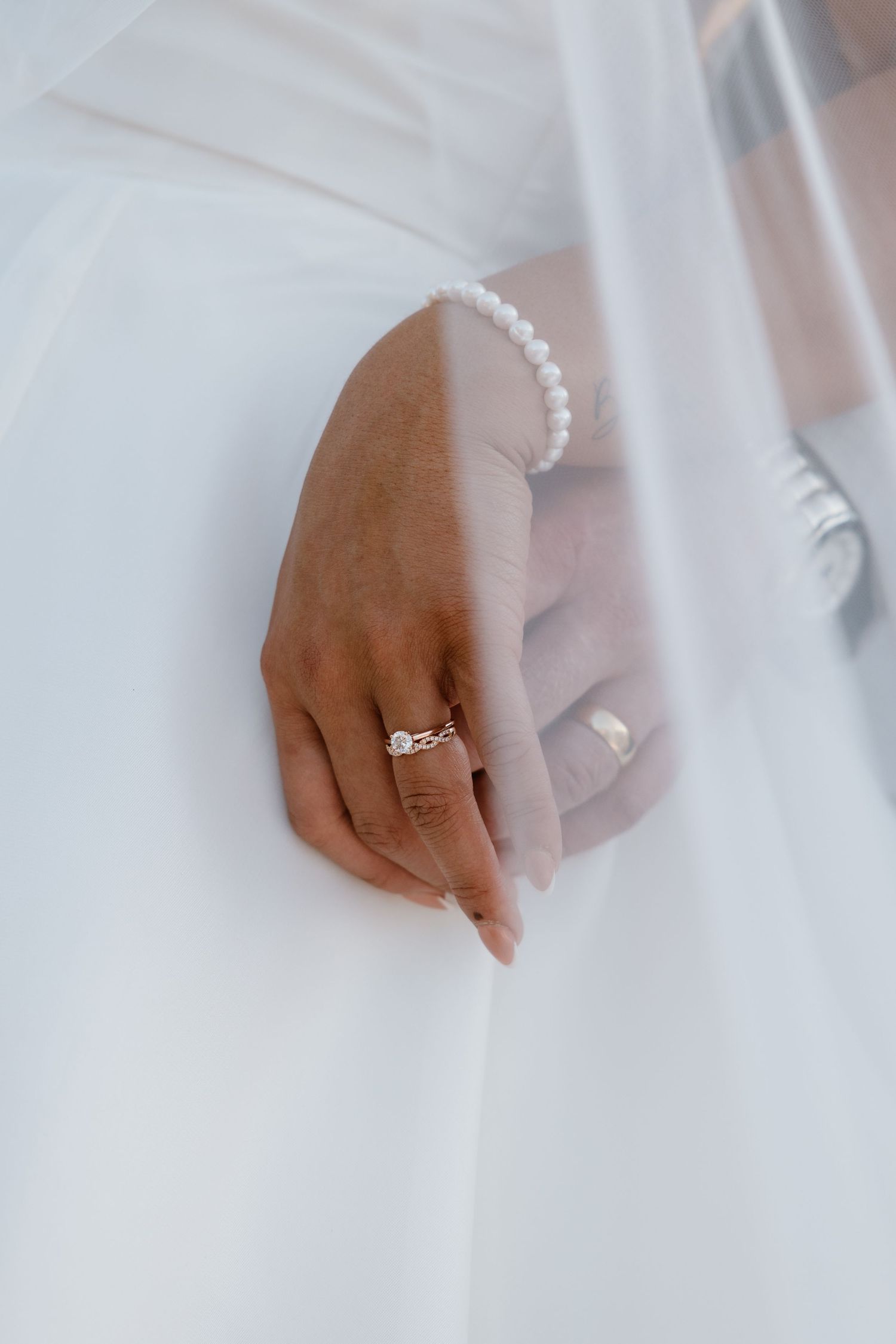 Close-up view of hands wearing a delicate wedding ring against a soft white wedding dress background.