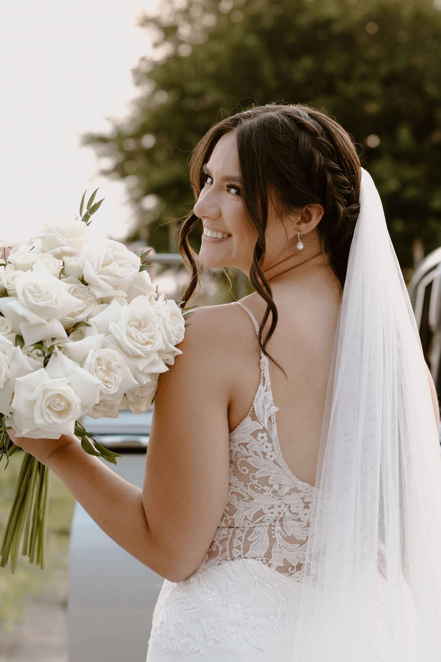 A bride in a white lace wedding dress holds a bouquet of white roses while wearing a flowing veil.