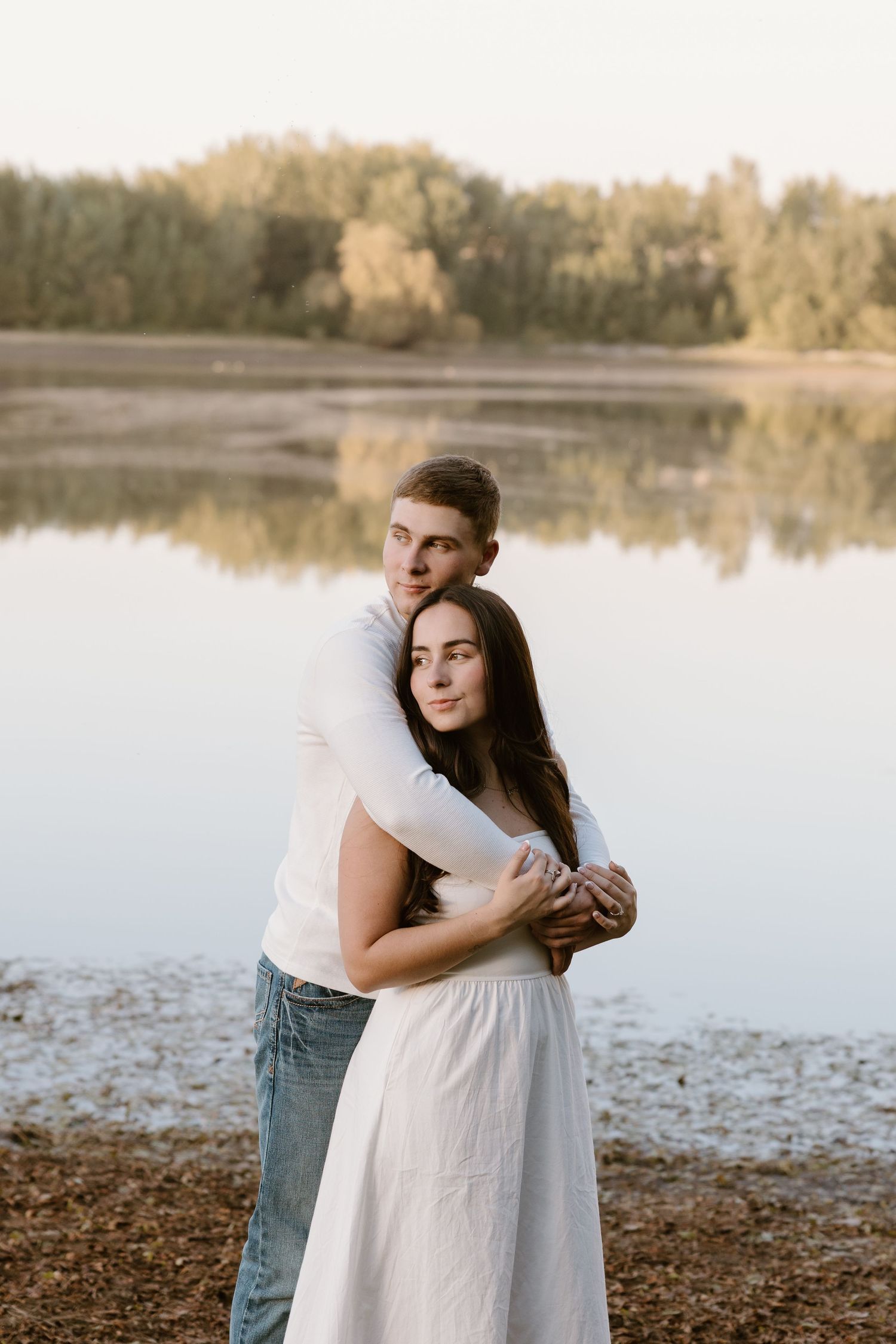 A couple embraces by a serene lake at sunset with warm golden light filtering through the trees.