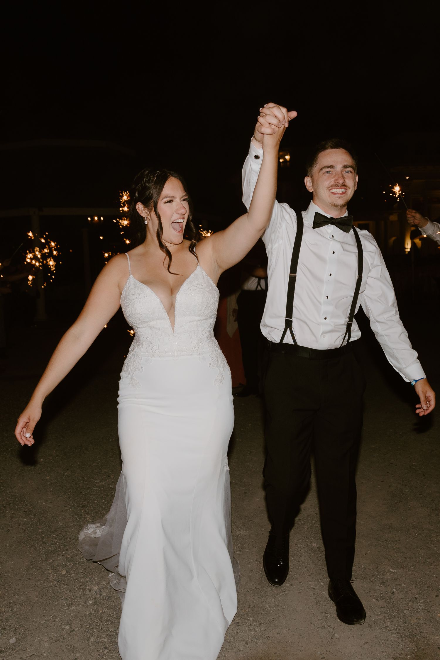A couple dances joyfully at their wedding reception, with the bride in white and groom in black with suspenders.