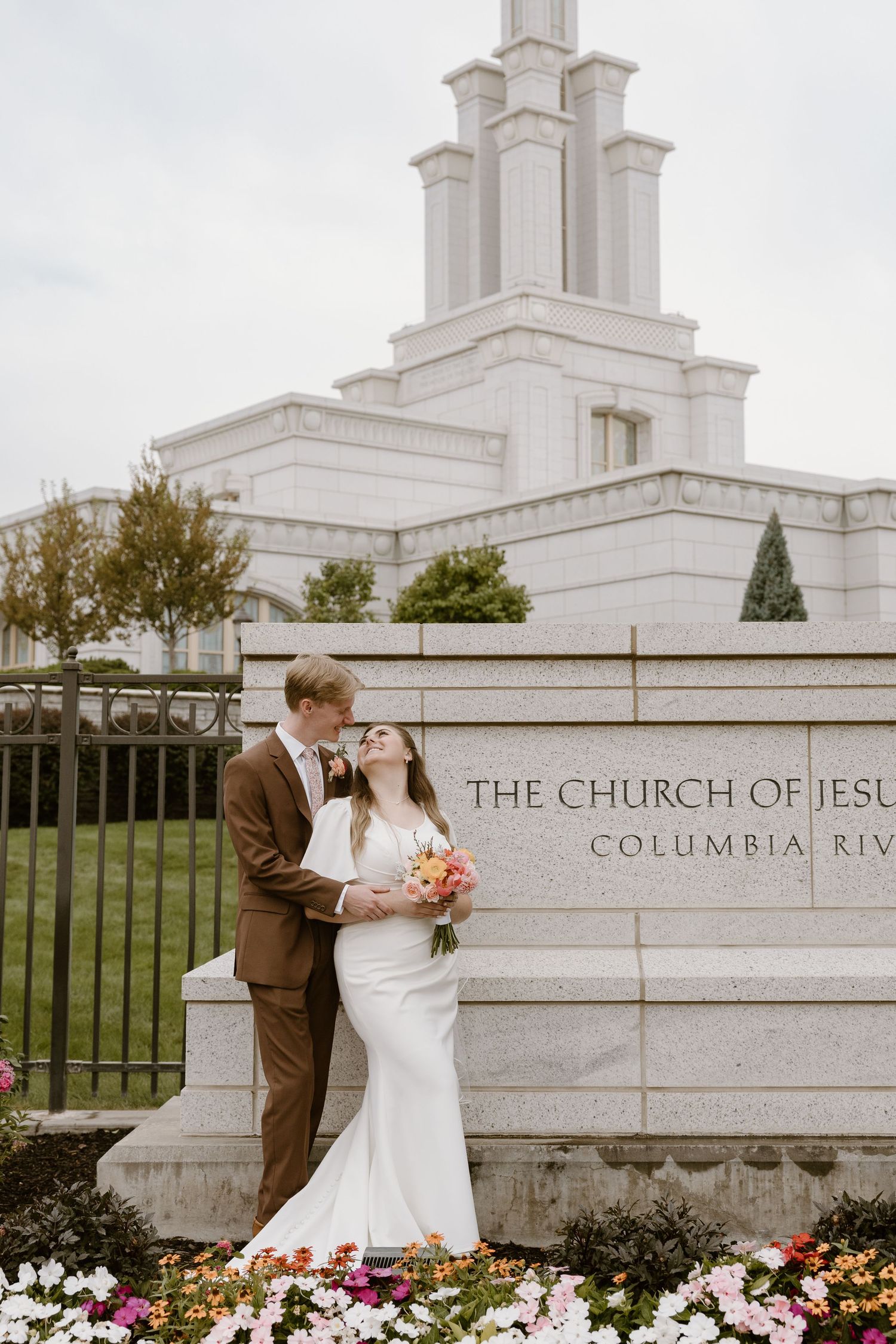 Wedding couple poses in front of white stone church building with floral landscaping.