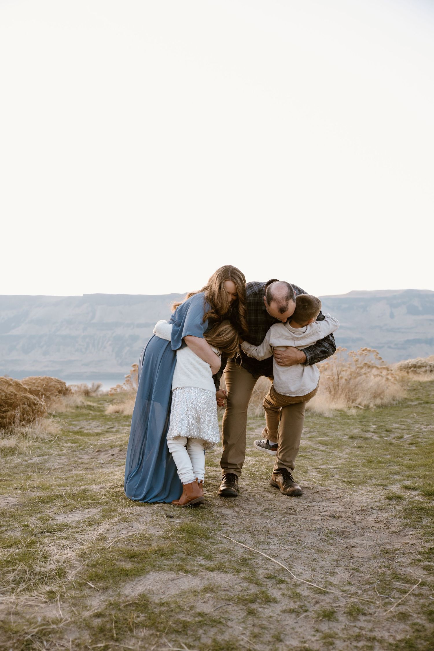 A father and mother bends down to hug their children on a rocky hillside against a misty landscape.