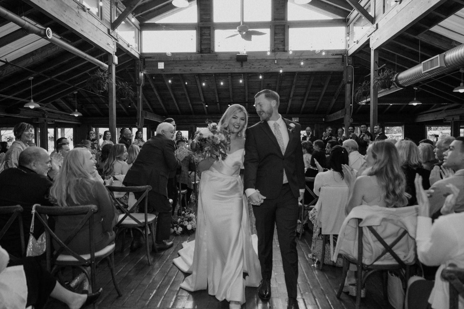 A black and white photograph of a couple sharing their first dance in a rustic wooden barn venue surrounded by guests.