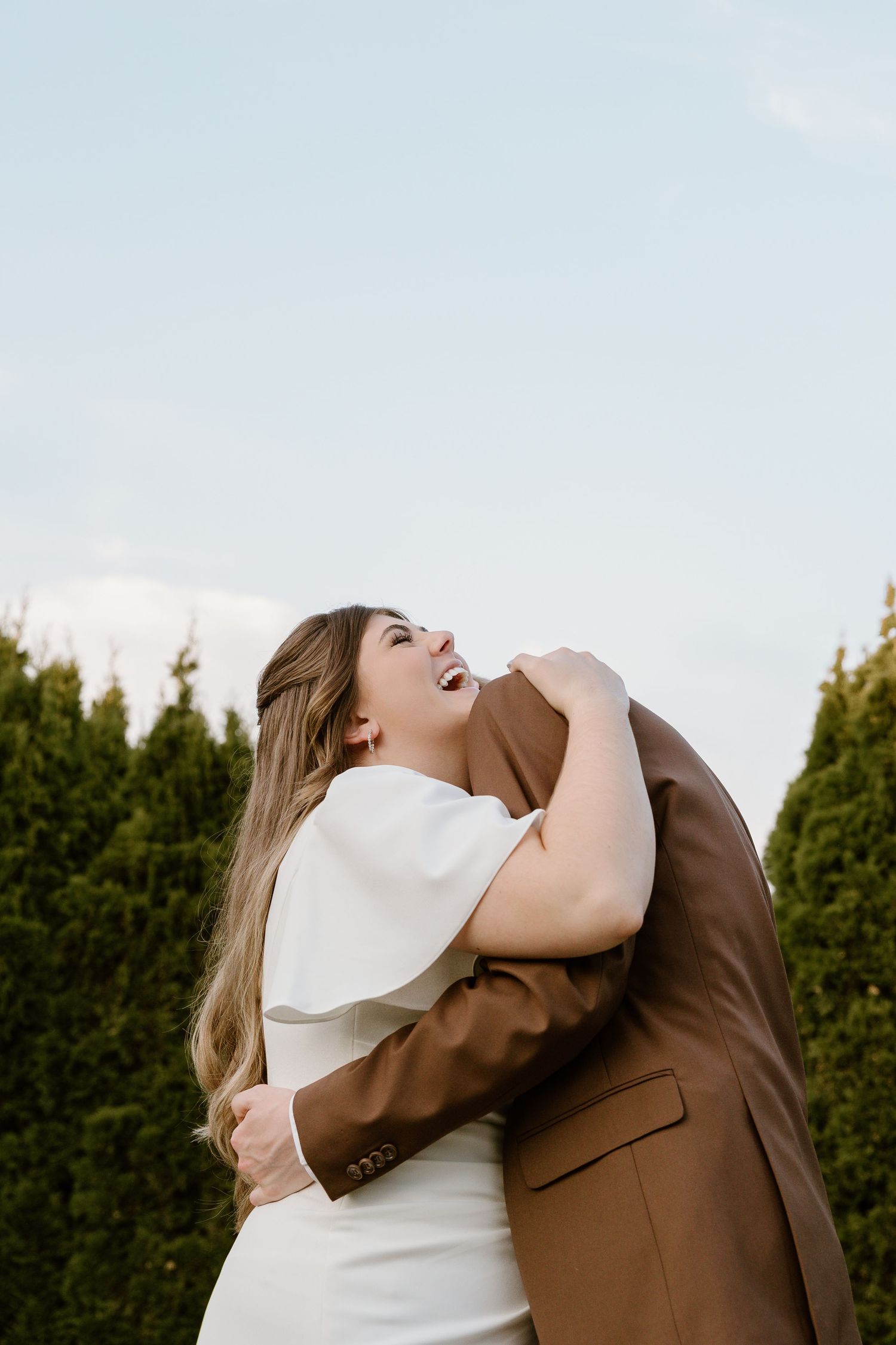 A joyful moment between newlyweds sharing laughter in a garden setting with evergreen hedges.