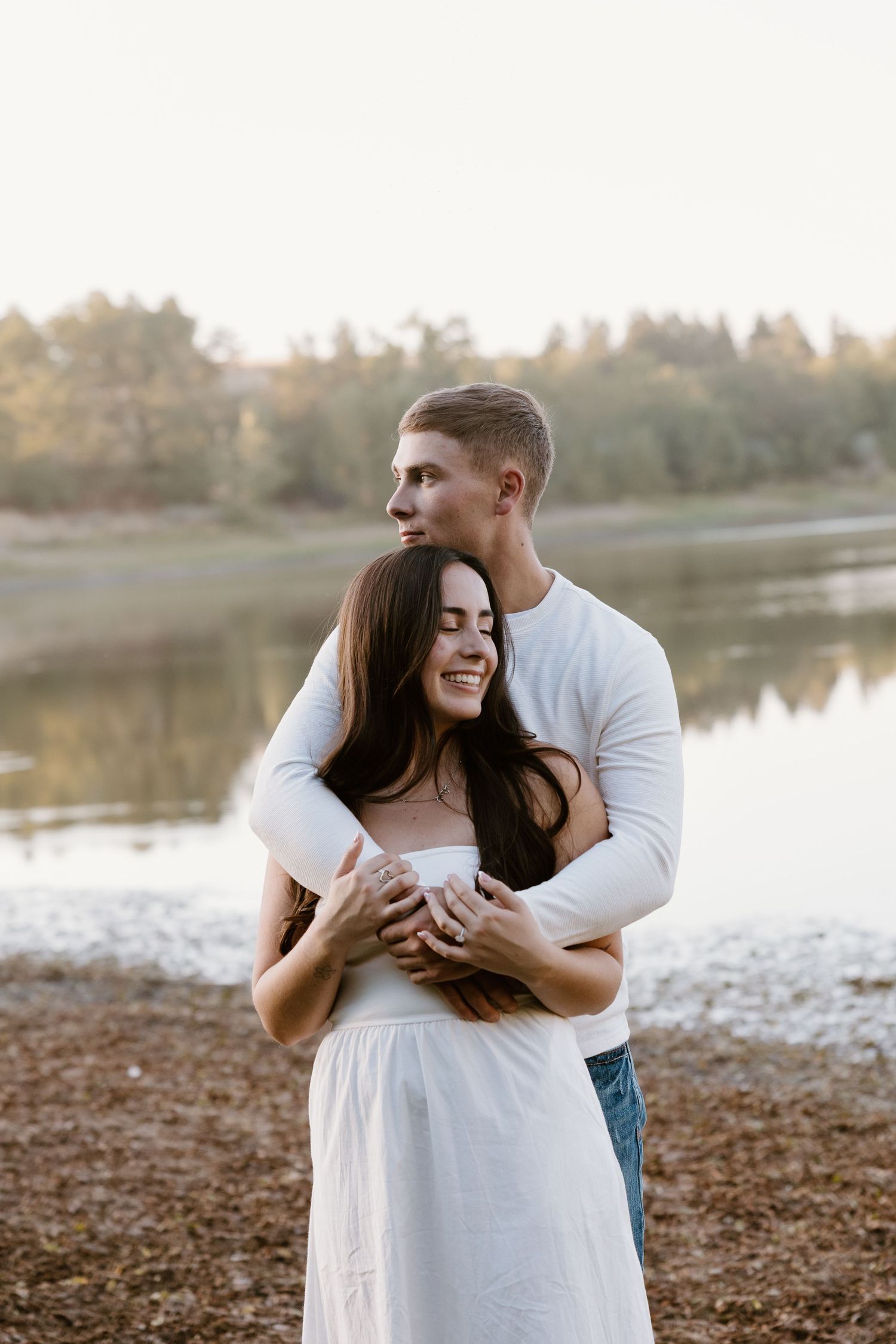 A couple embraces lovingly by a serene lake at sunset during their engagement photoshoot.