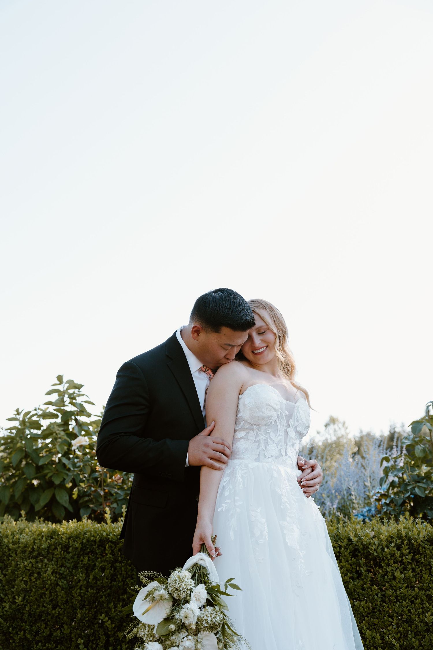 Romantic wedding couple embraces in a garden setting with greenery and white flowers at sunset.