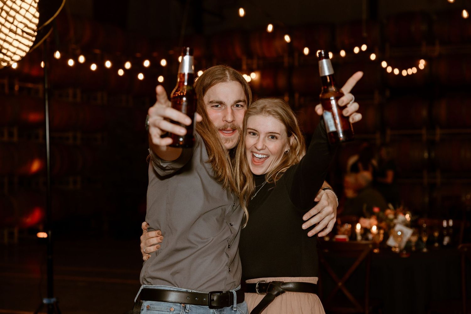 A couple celebrates at night under string lights, raising sparklers and embracing while smiling at the camera.