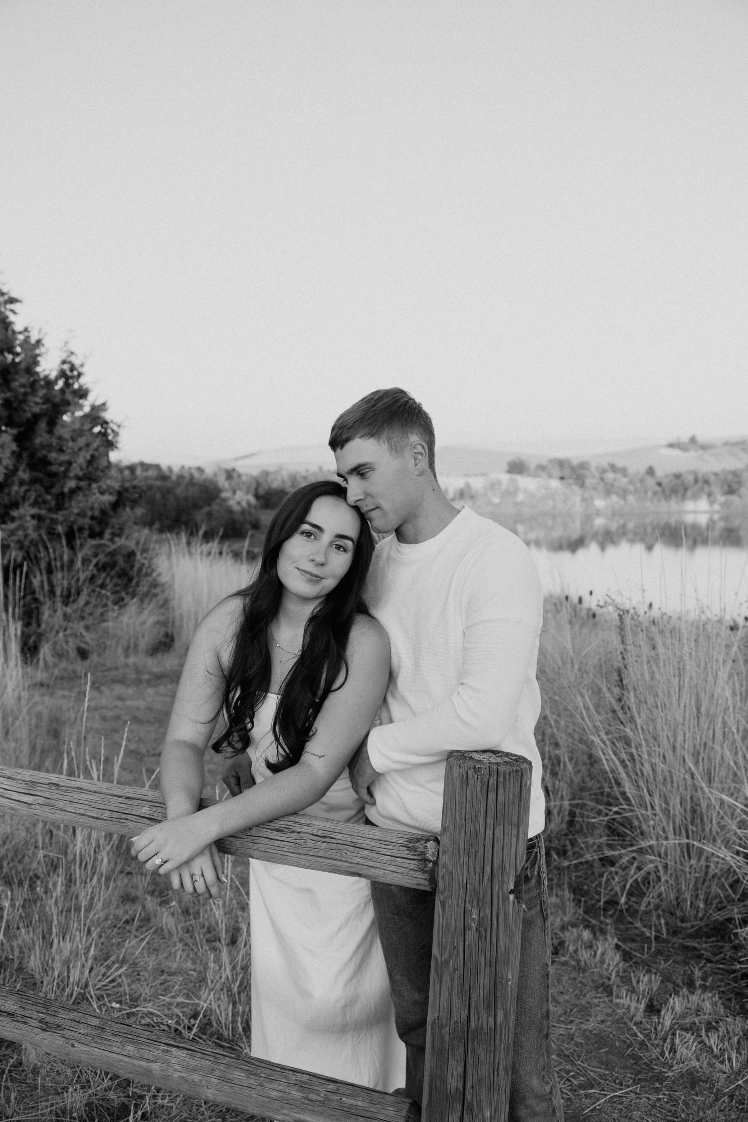 A couple shares romantic moments along a rustic wooden fence in a field during a black and white photoshoot.
