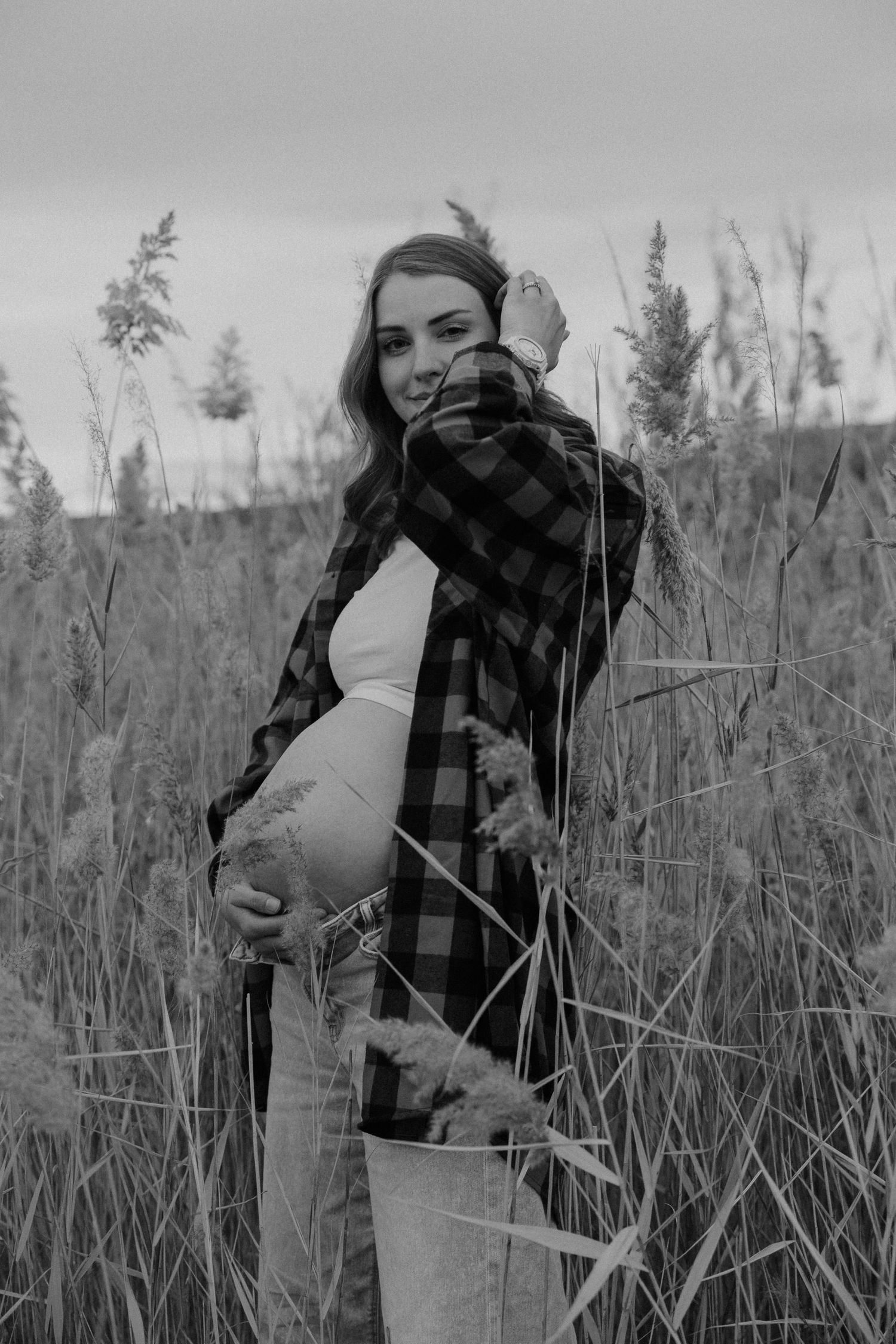 Black and white maternity photoshoot in tall grass field with plaid shirt and natural outdoor setting.