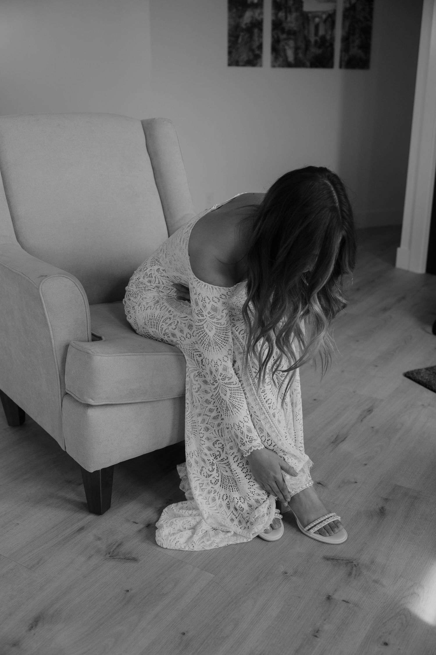 A bride in a white lace wedding dress adjusts her shoes while sitting on a light colored chair.