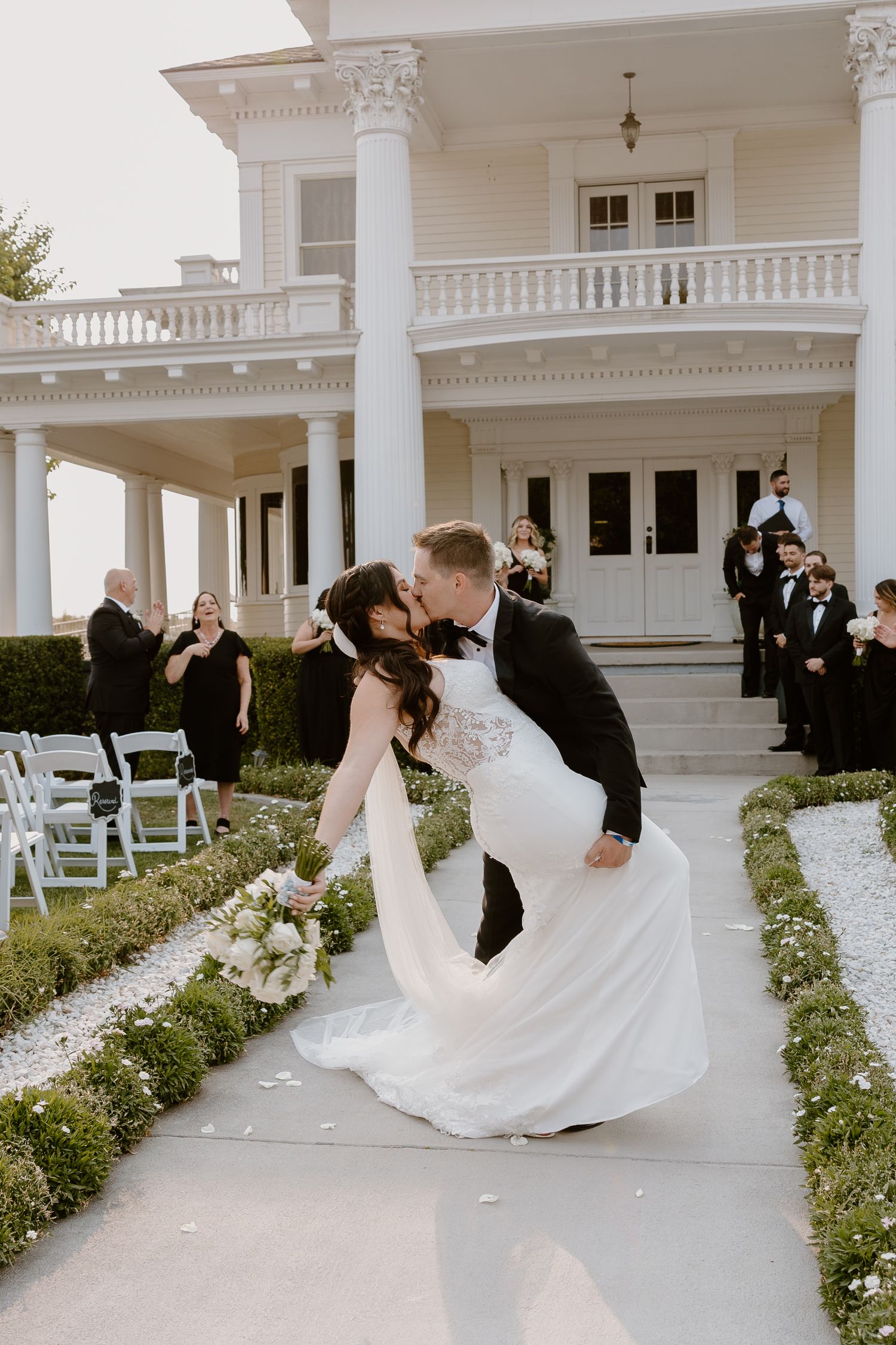 Elegant wedding couple dips in romantic dance sequence on white mansion porch with columns and garden backdrop.