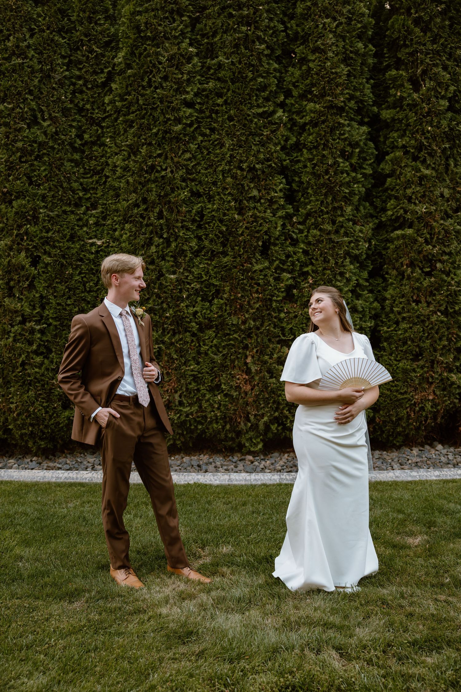 A couple in wedding attire shares playful moments on grass in front of a green hedge backdrop.