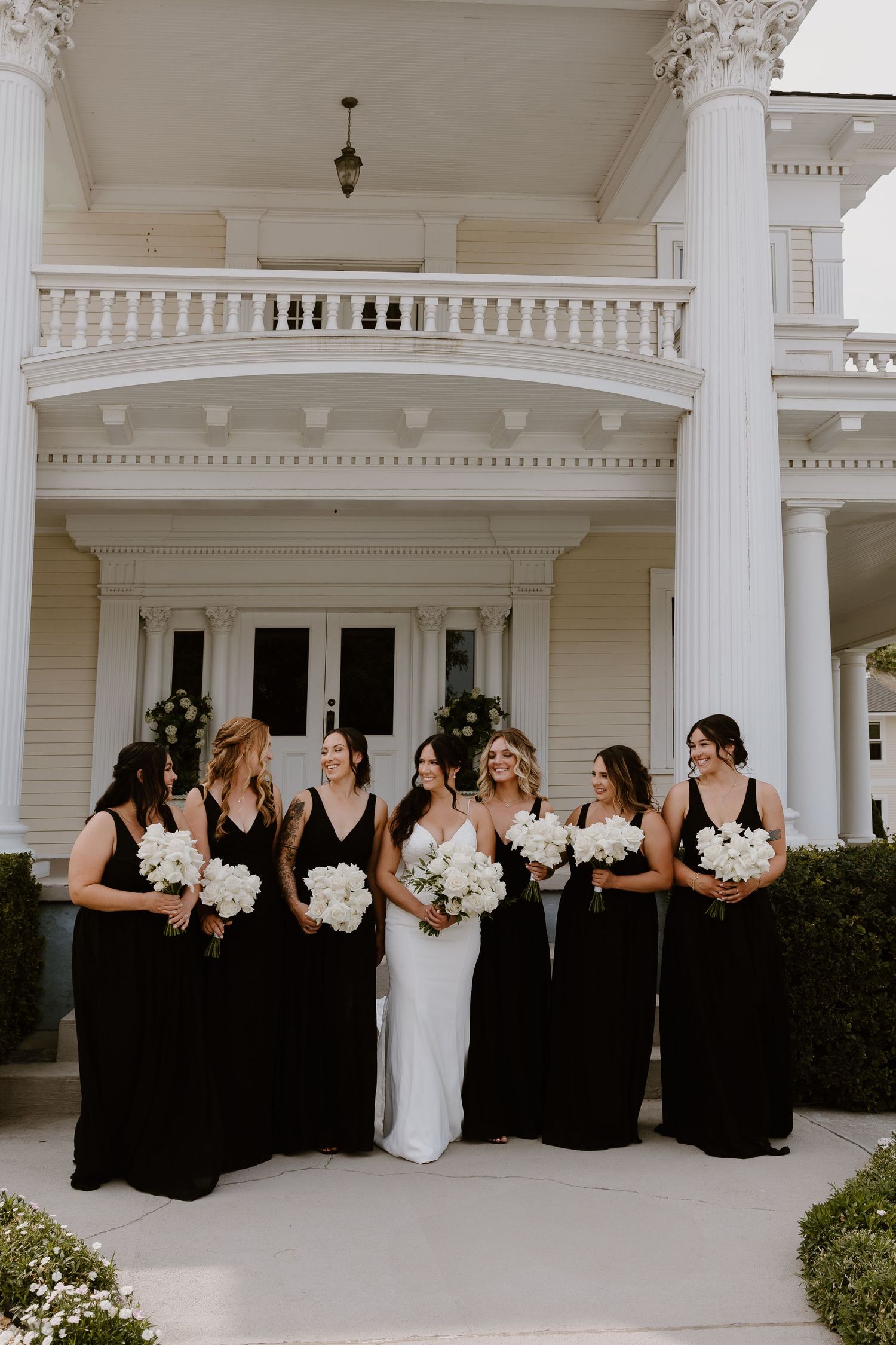 Bridesmaids in elegant black dresses and one in white pose together on a grand white columned porch.