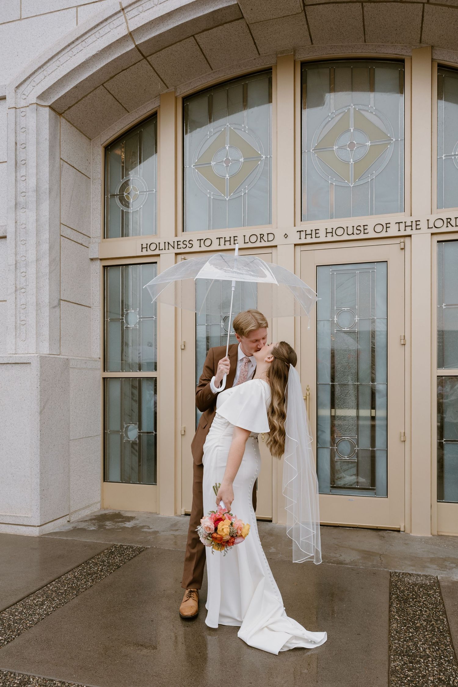 A romantic wedding couple shares a tender kiss in front of a church building with ornate architectural details.