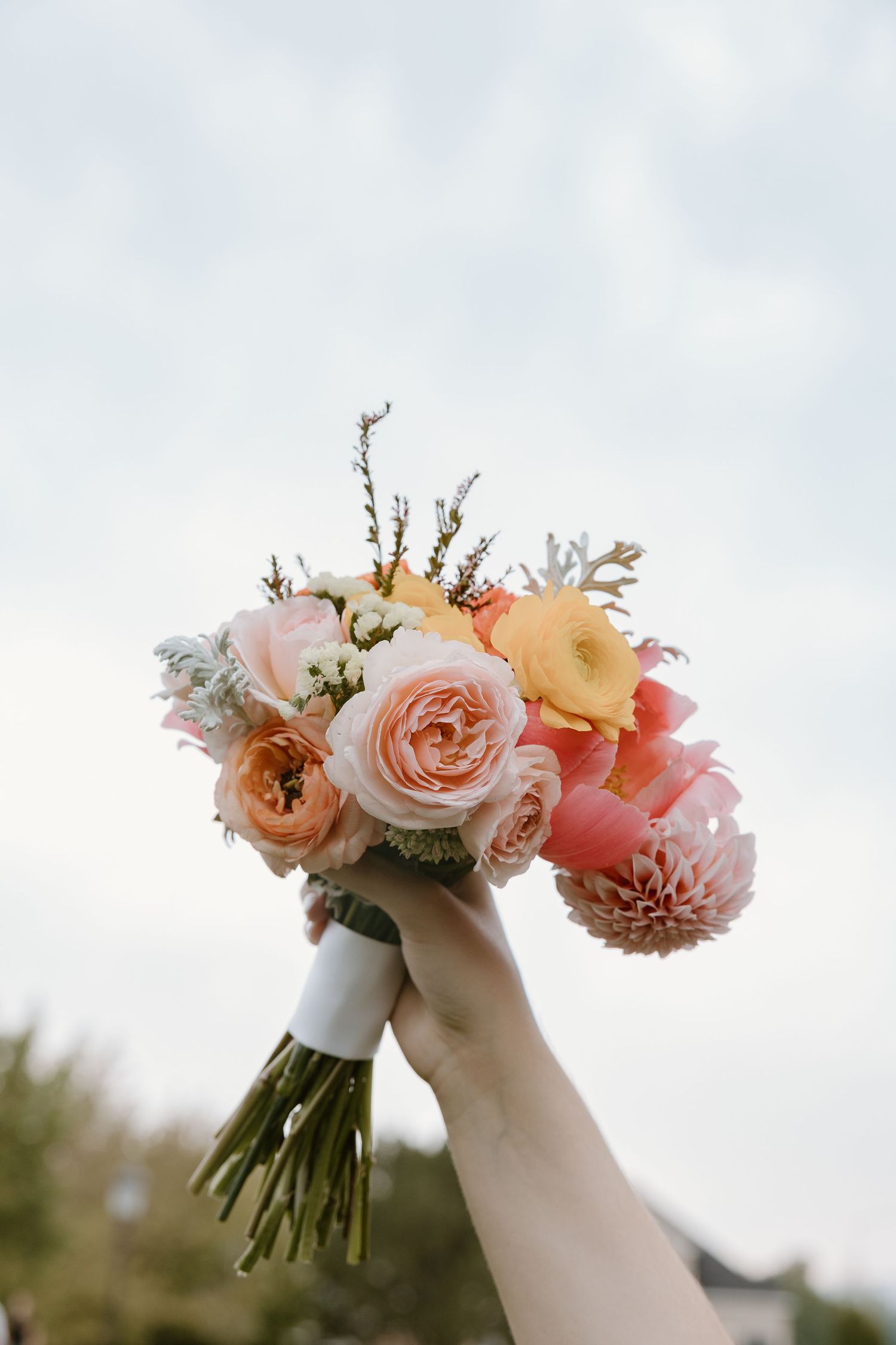 A hand holds up a romantic peach and coral wedding bouquet with roses and dahlias against a soft cloudy sky.