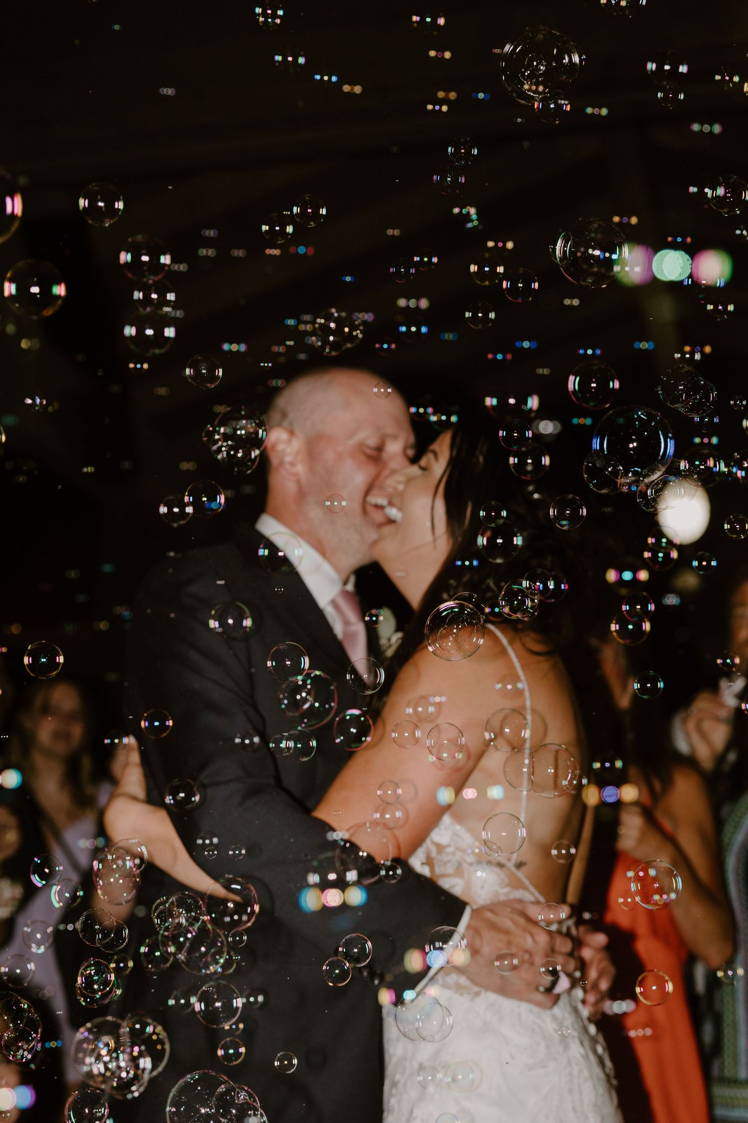 A romantic wedding couple shares their first dance surrounded by floating bubbles in a dimly lit reception space.