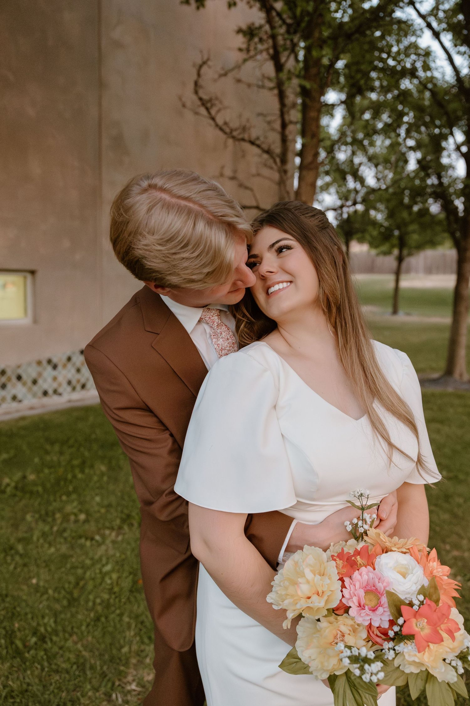 A couple shares an intimate moment in their wedding attire while holding a coral and peach floral bouquet.