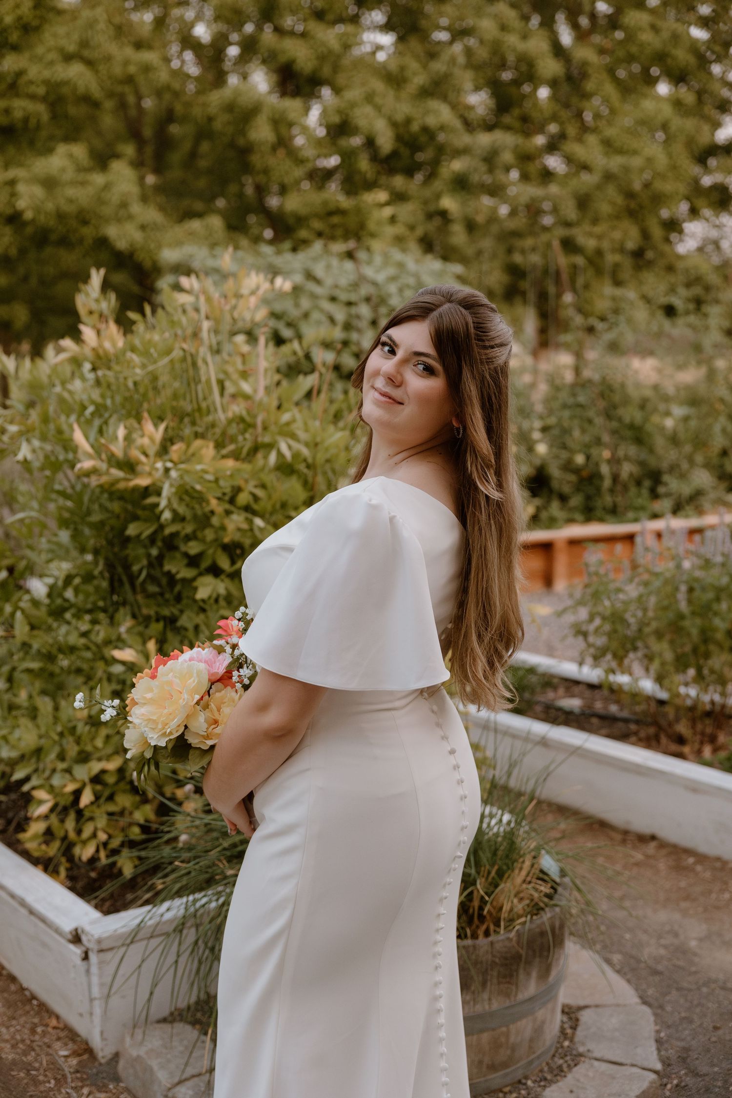 A person in an elegant white wedding dress poses in a garden setting holding a bouquet of flowers.