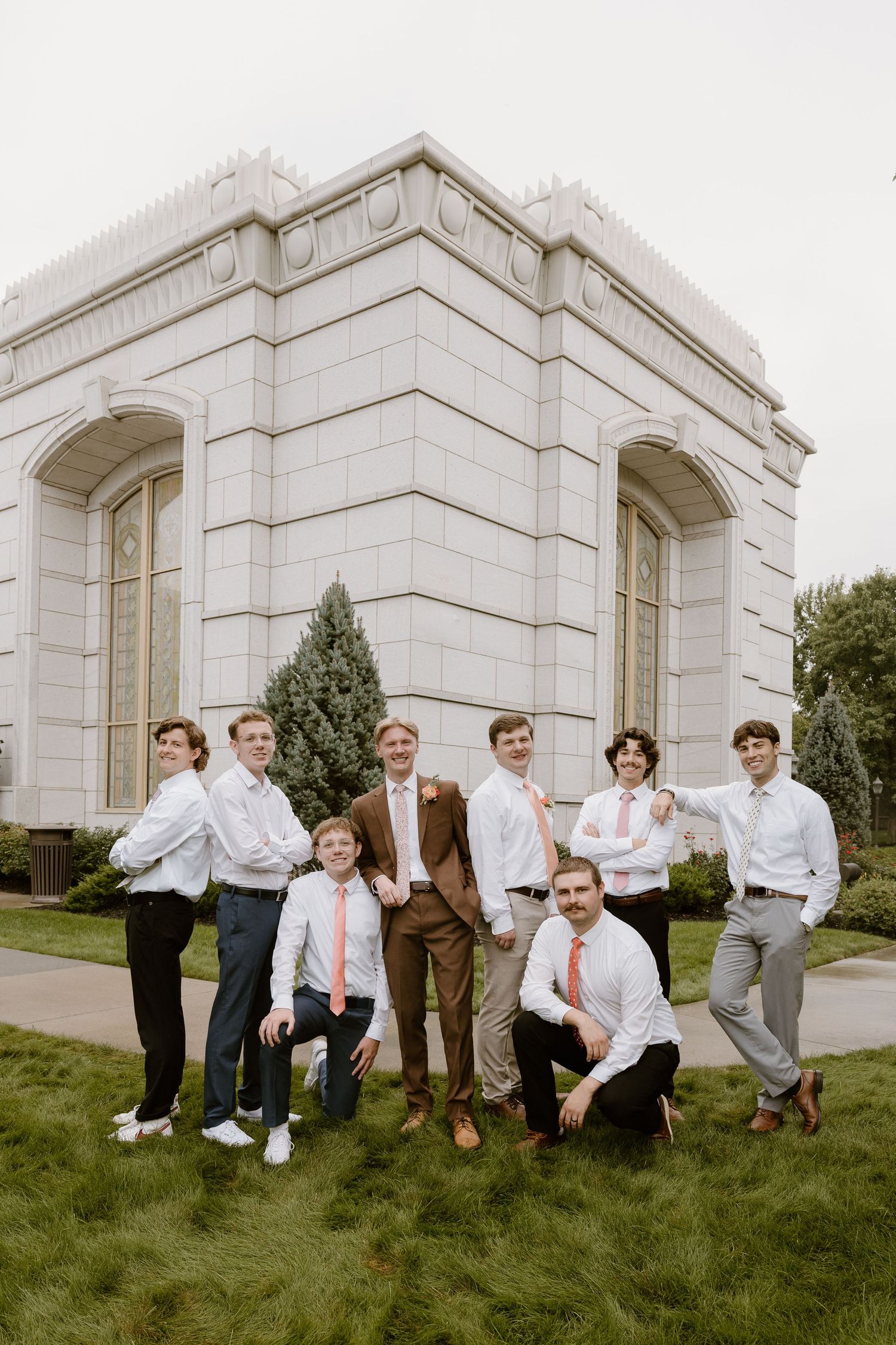 Groomsmen strike playful poses while sitting and kneeling on grass in front of a stone building.