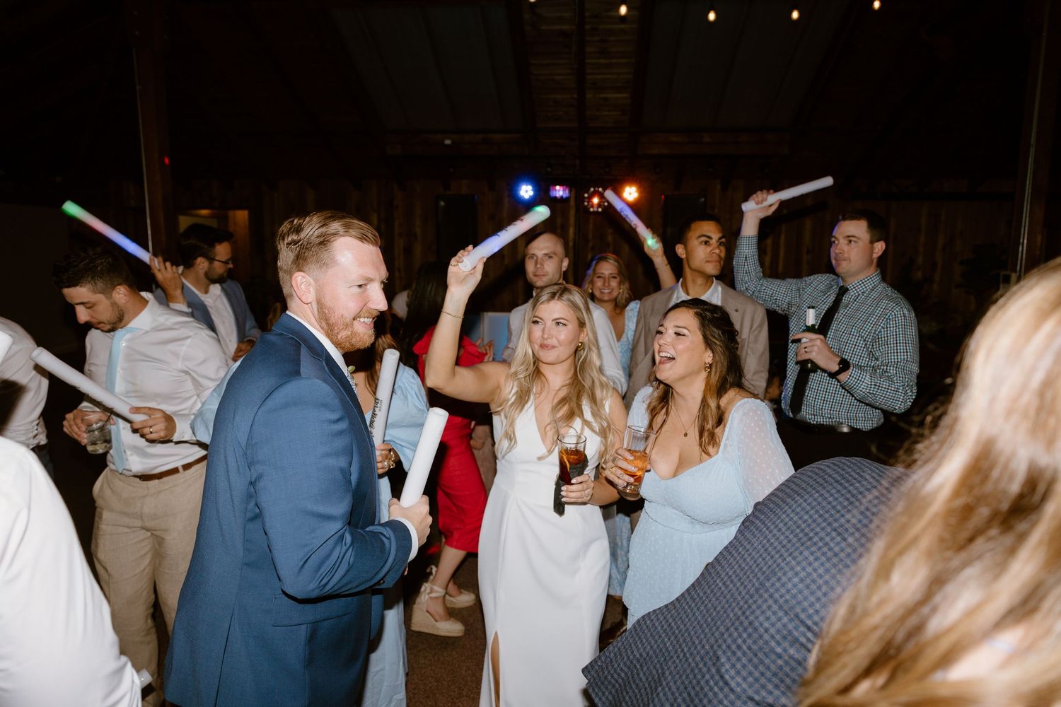Wedding guests dance while holding illuminated LED foam sticks on a dark dance floor at a reception celebration.