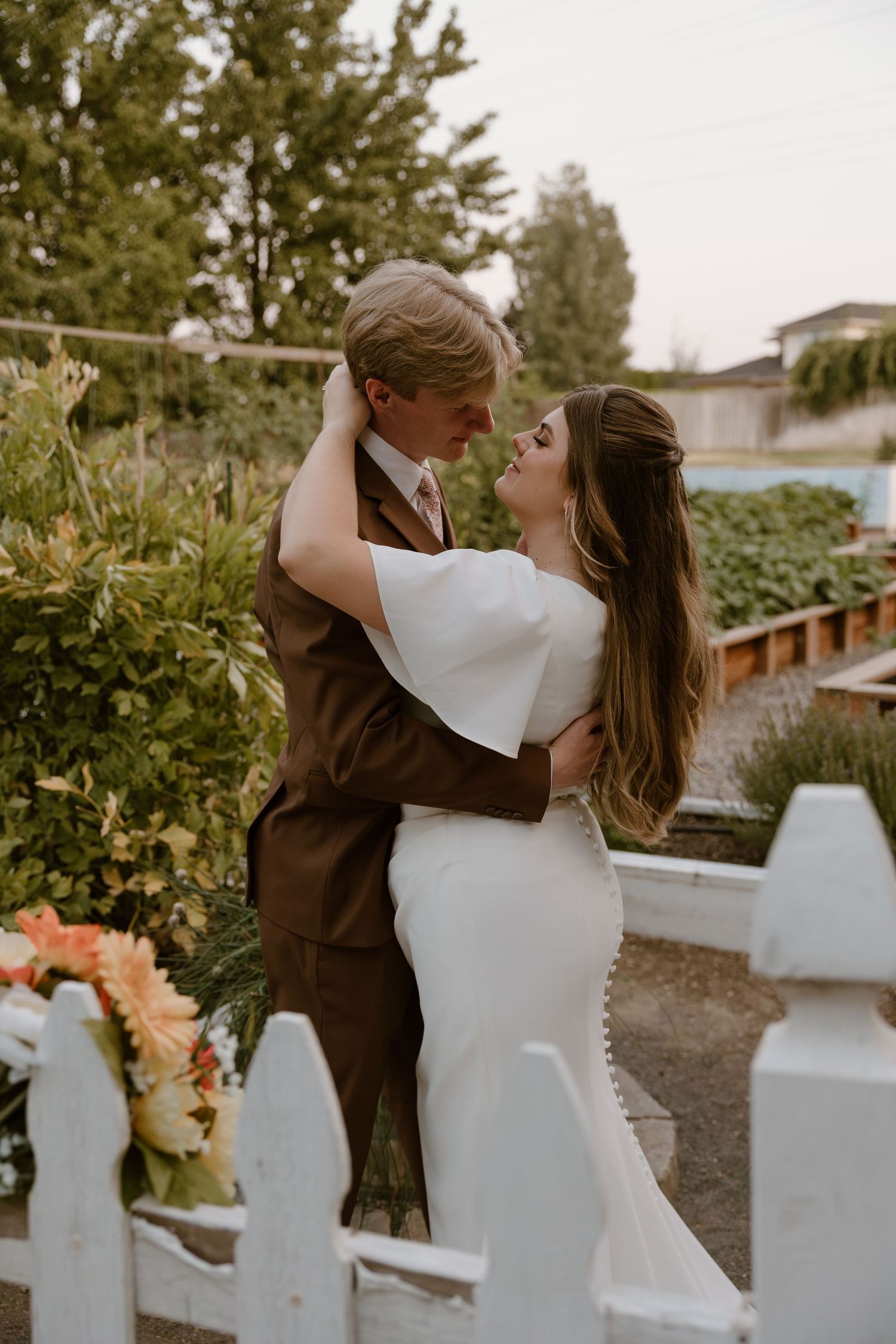 A romantic wedding couple embraces by a white picket fence in a garden setting at sunset.