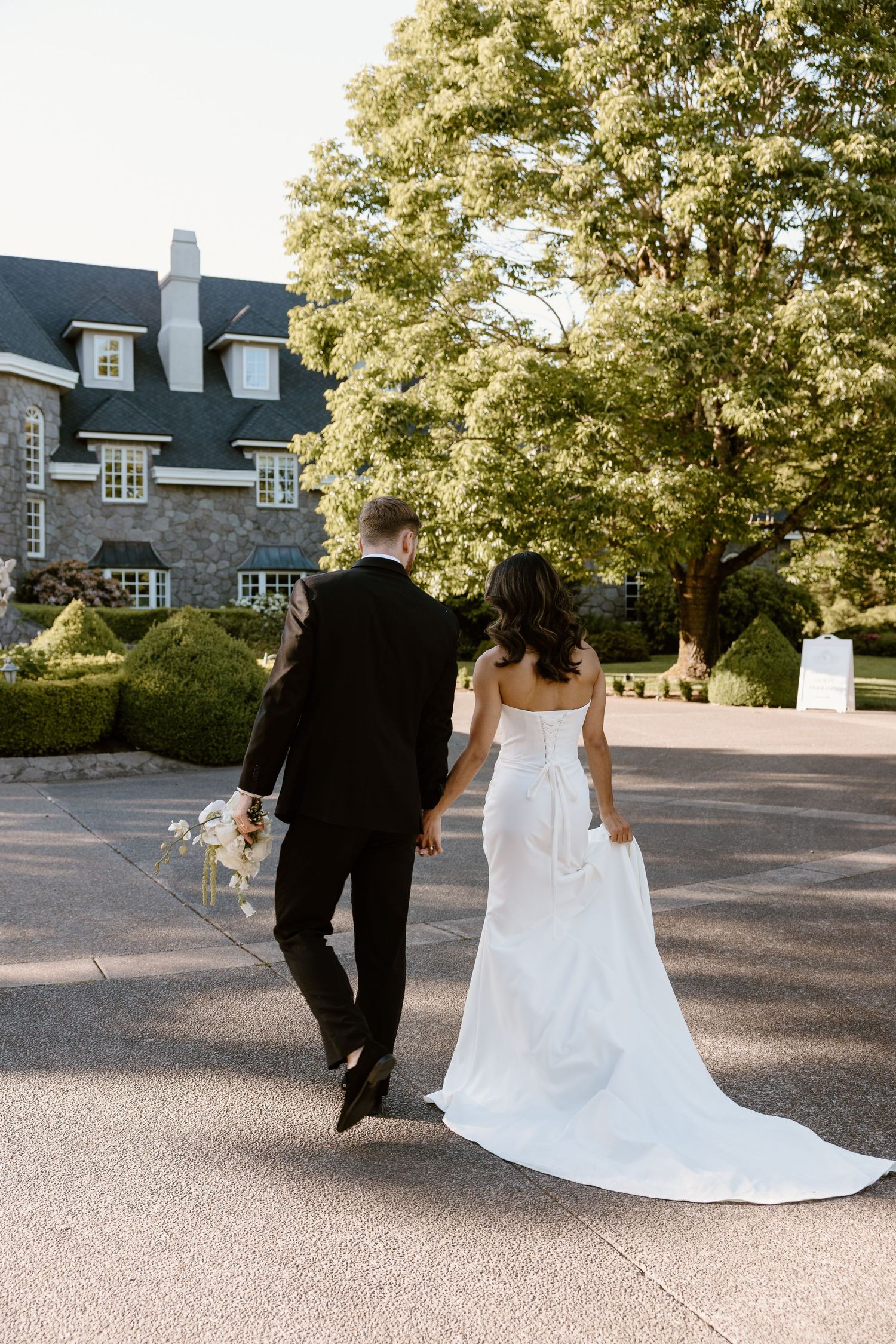 A couple in wedding attire walk away as husband and wife together on a paved outdoor area with trees in the background.