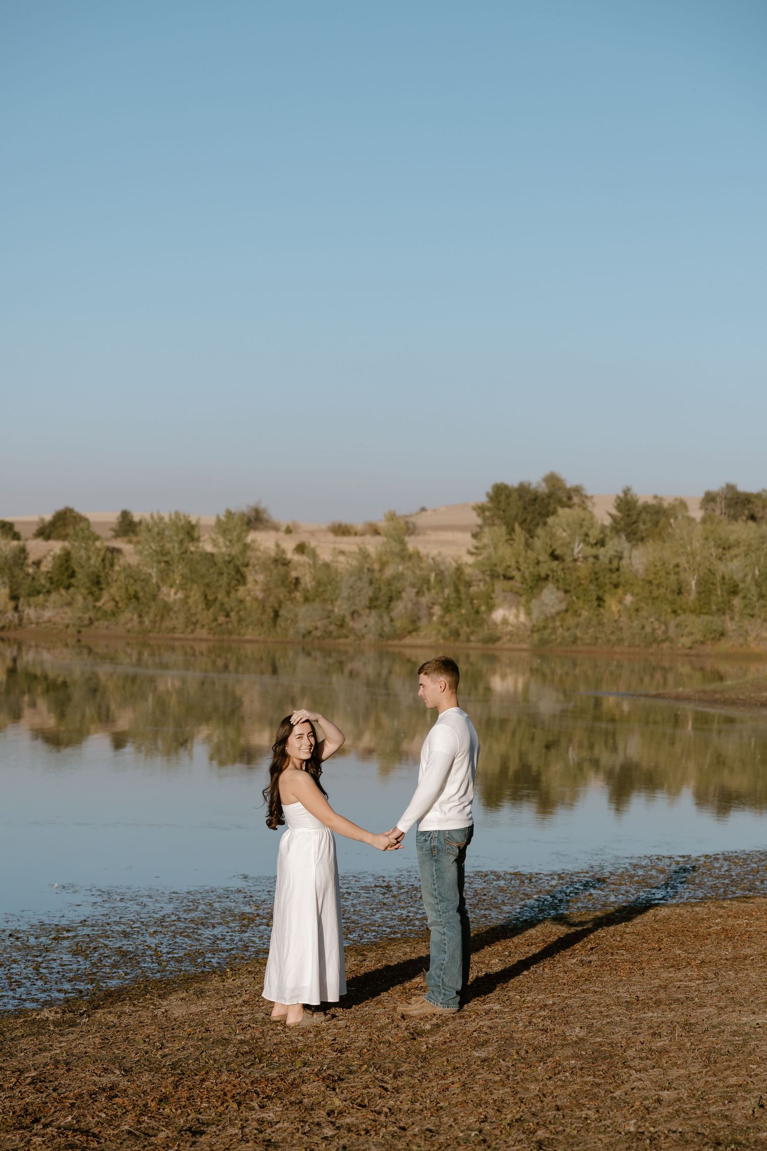 A couple in formal attire stands together on a lakeside shore during a romantic outdoor photoshoot at sunset.