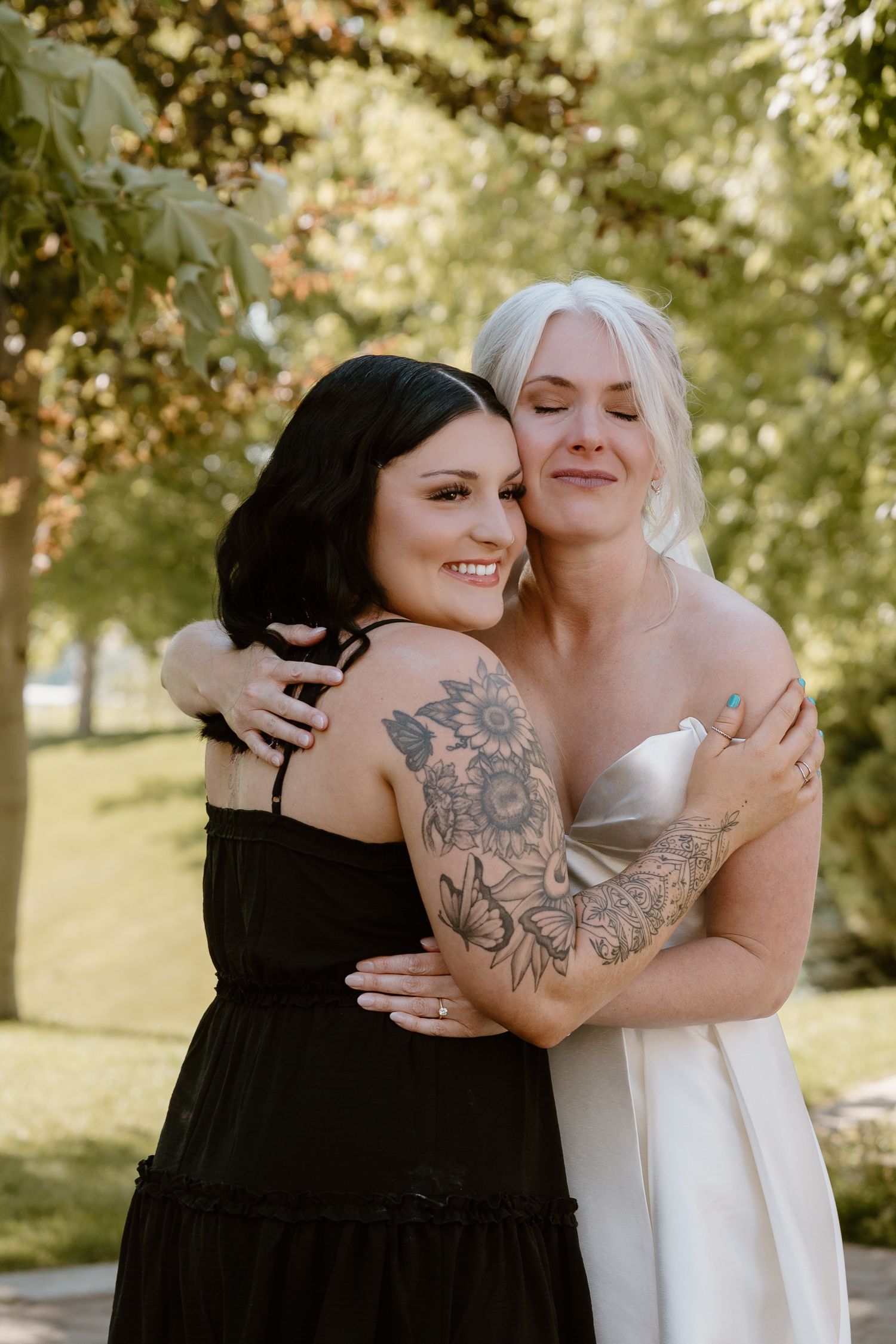 Two people sharing a warm embrace outdoors in a sunlit garden setting, one wearing black and one wearing white.