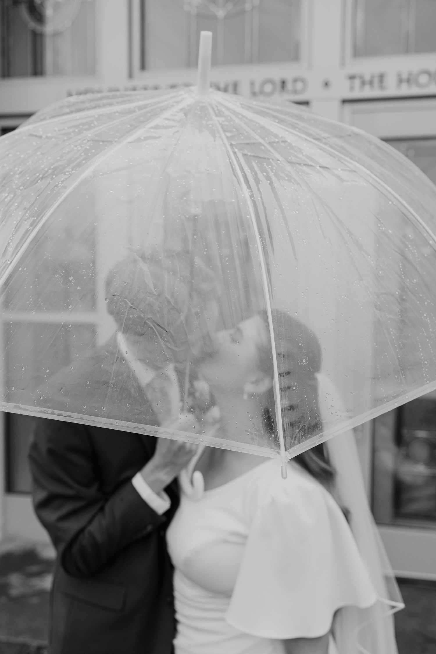 A couple shares a romantic kiss under a clear umbrella on a rainy day in black and white.