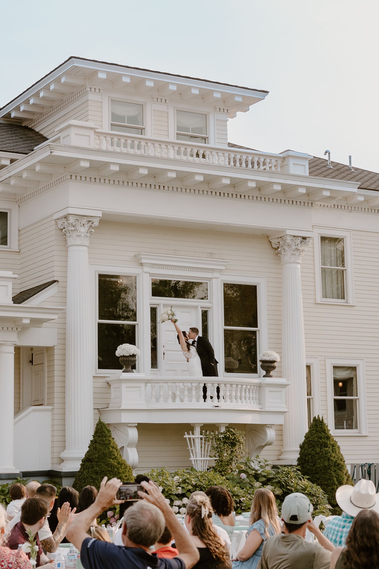 A crowd waves at a white Victorian-style house with ornate trim and columns during a celebratory gathering.