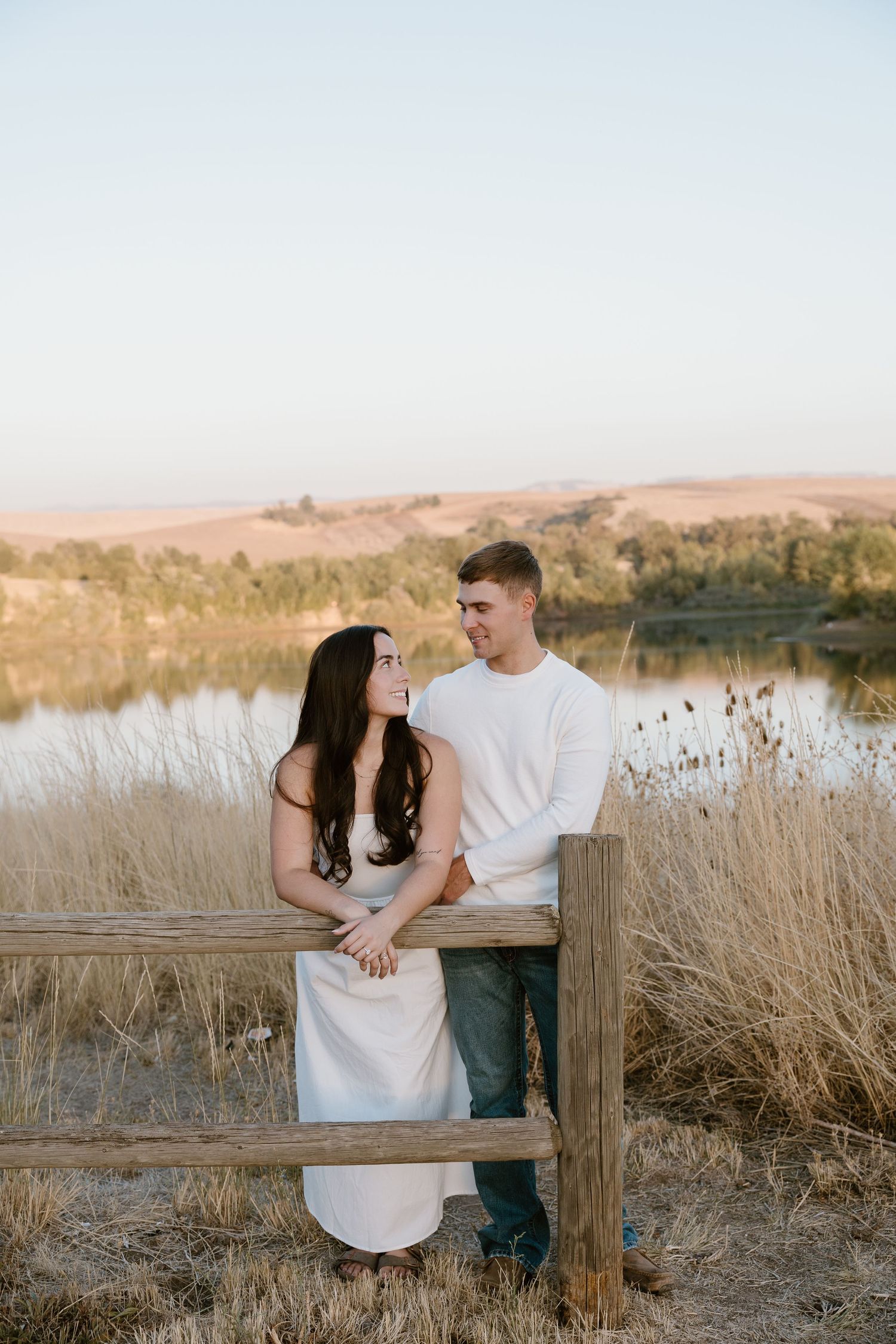 A romantic couple shares a tender moment by a wooden fence near a lake at sunset with golden grasses surrounding them.