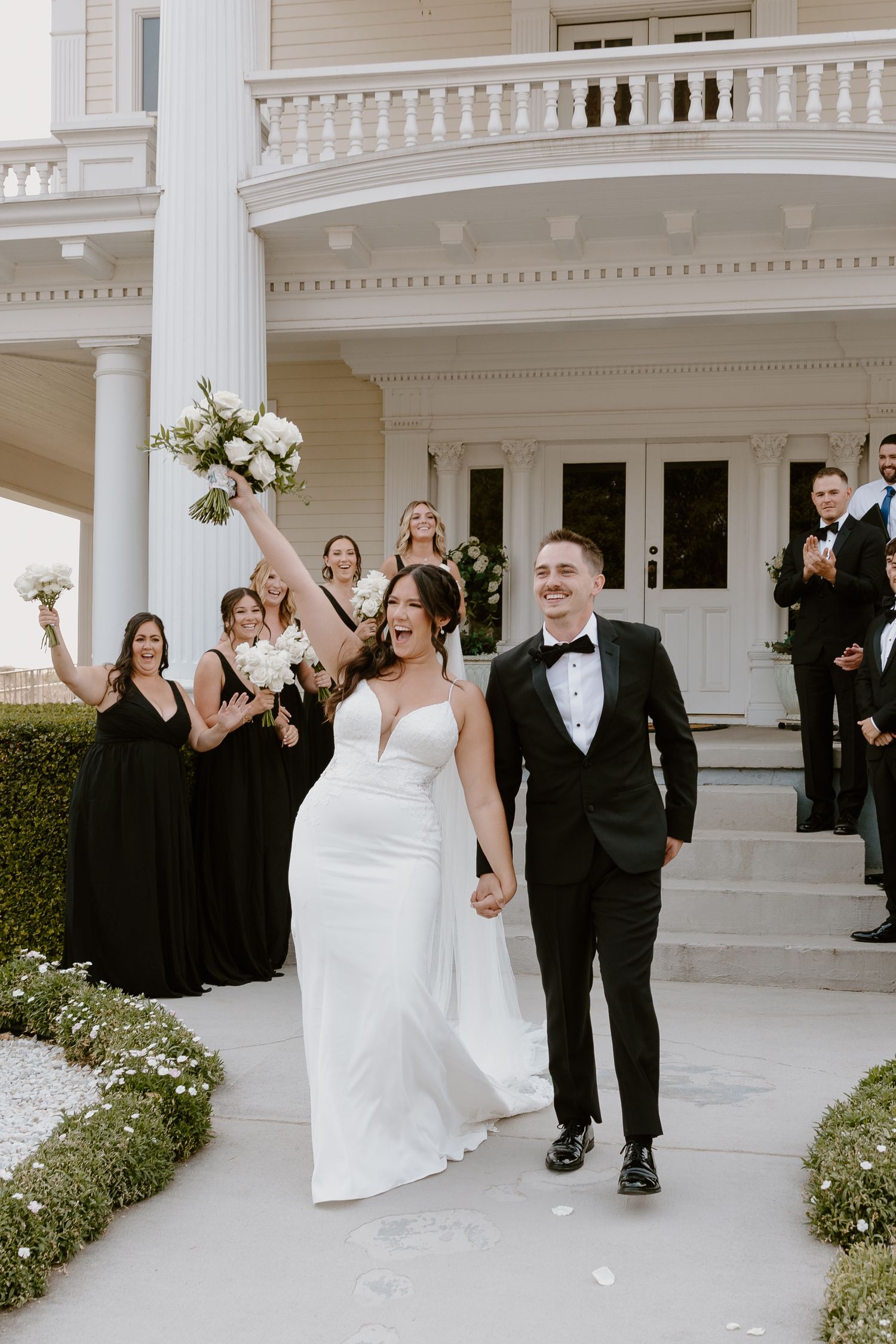 Wedding party celebrates joyfully outside elegant white columned mansion with ornate balustrades.