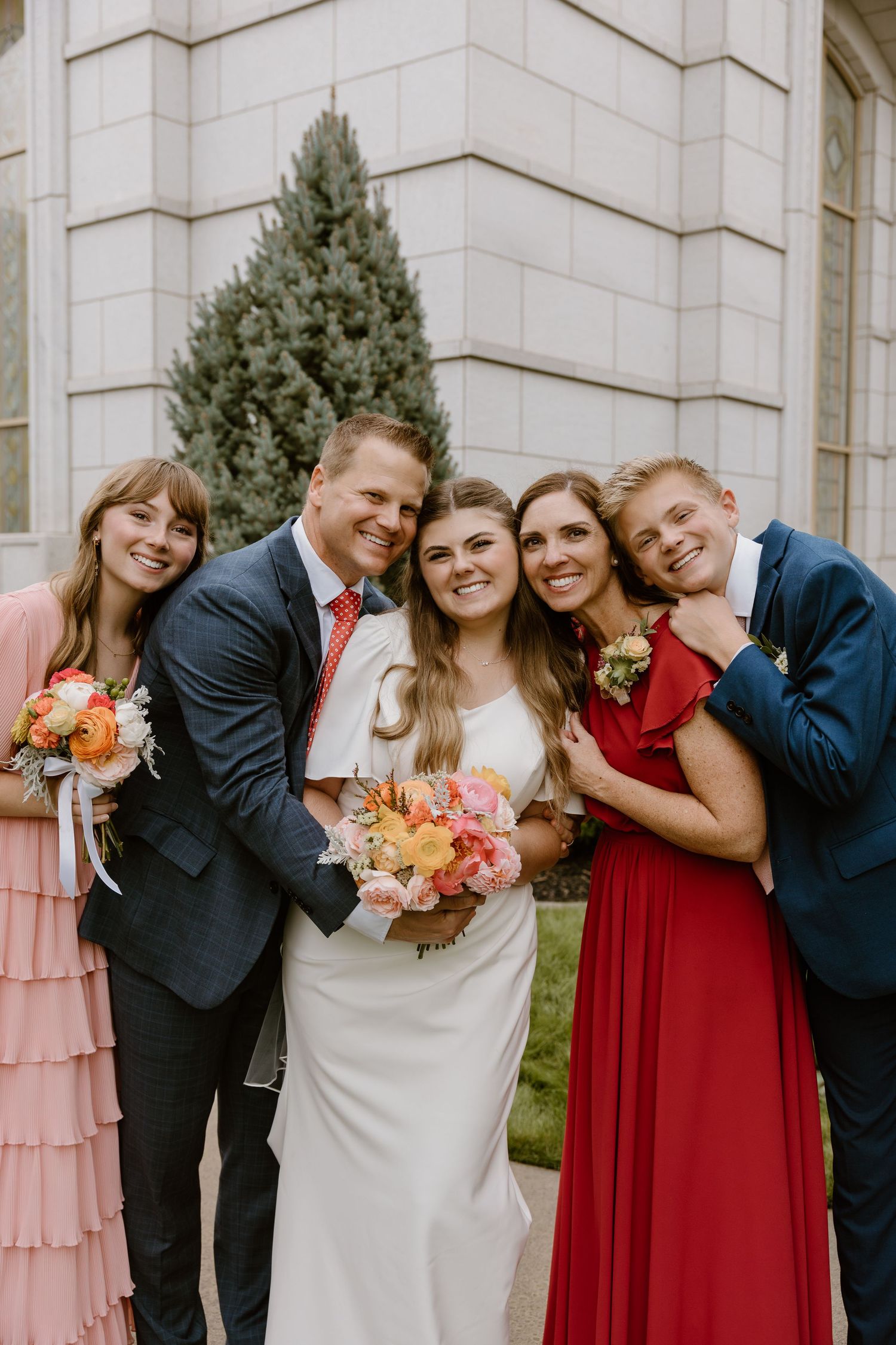 Wedding party in formal attire poses together against light stone building with bridesmaids in red and pink dresses.