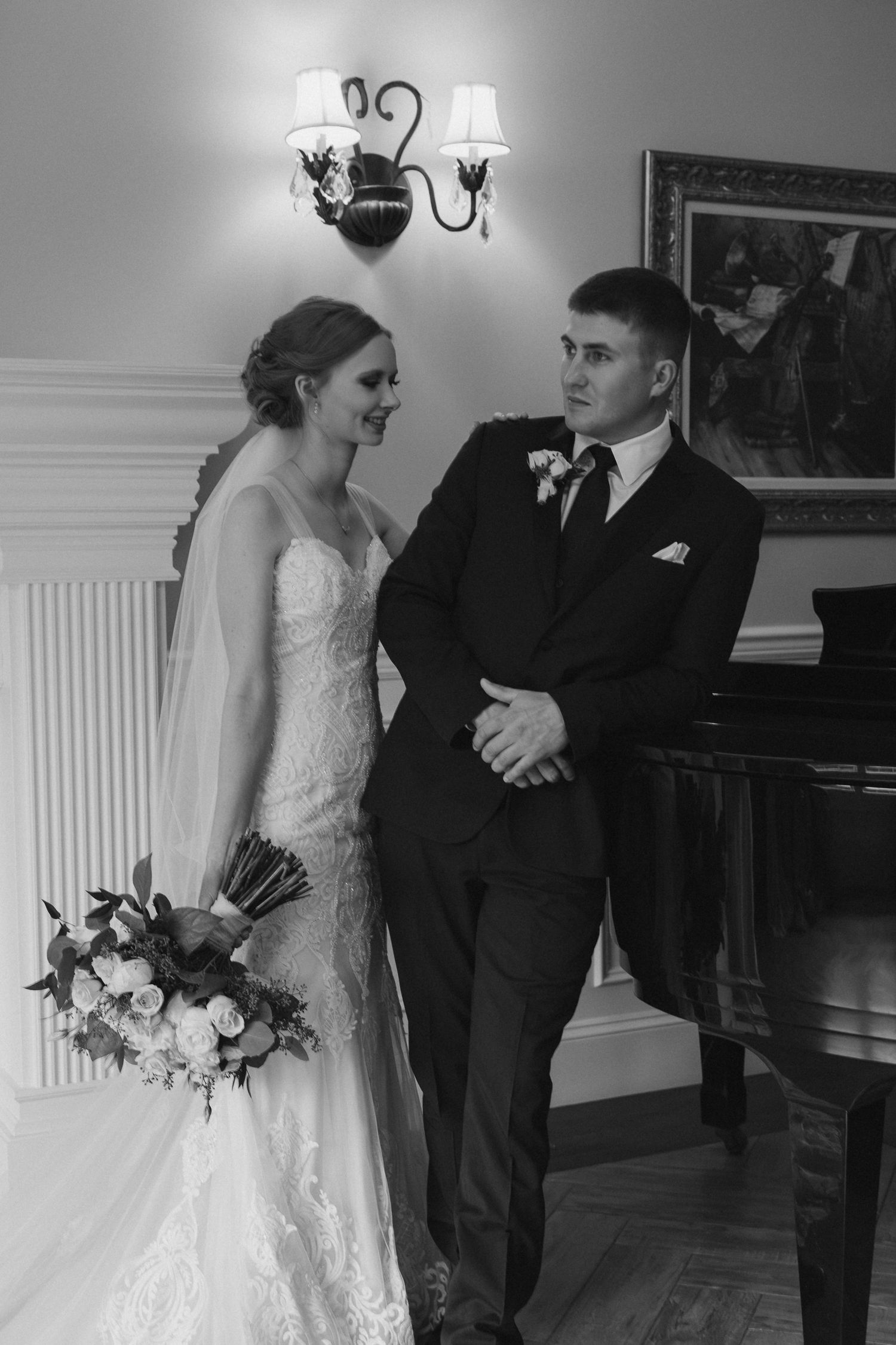 A black and white wedding photo showing a couple sharing a candid moment next to a piano in an elegant room.