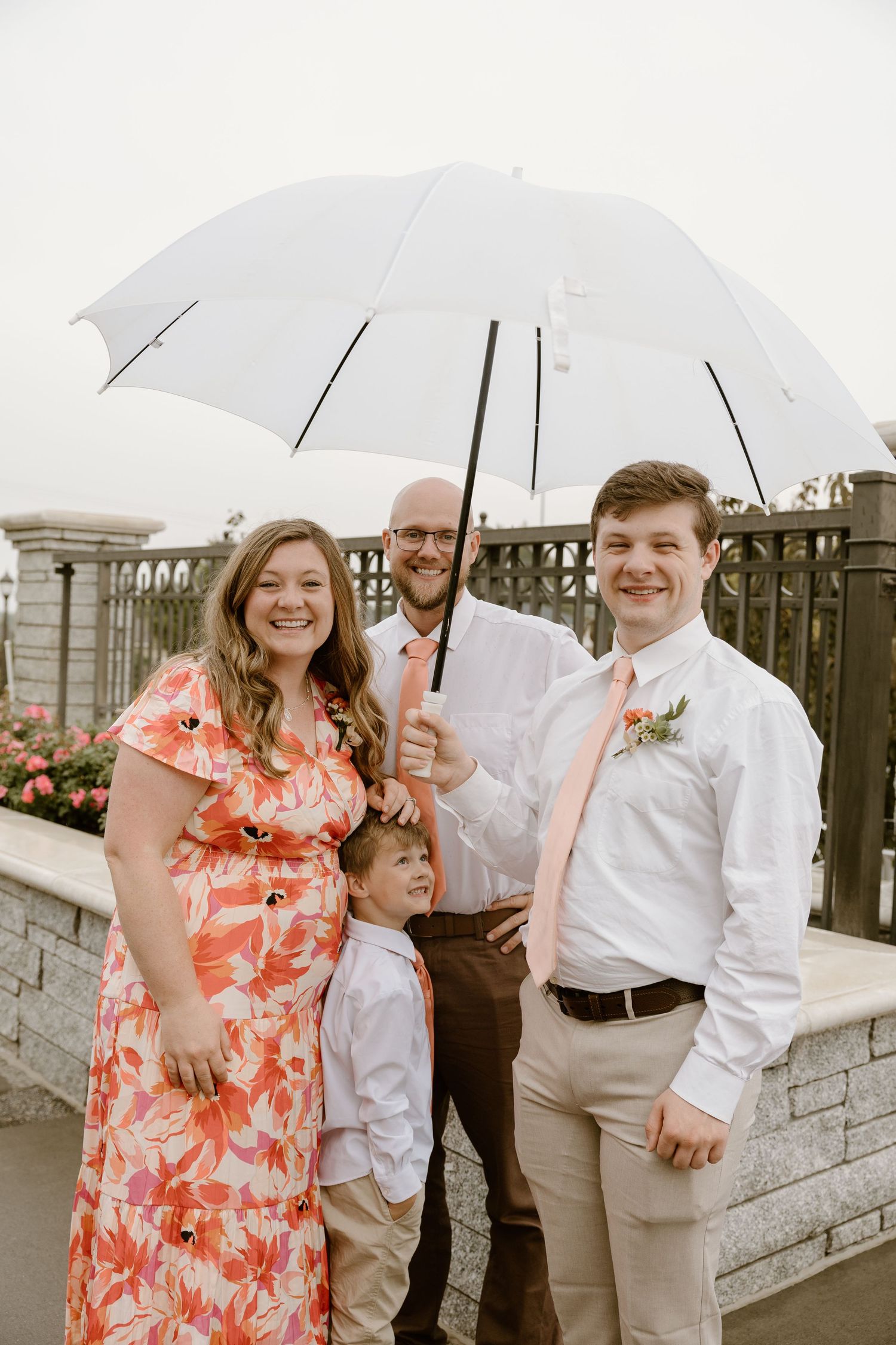 A group of people holding clear umbrellas pose together on a rainy day, with one wearing a coral floral dress.