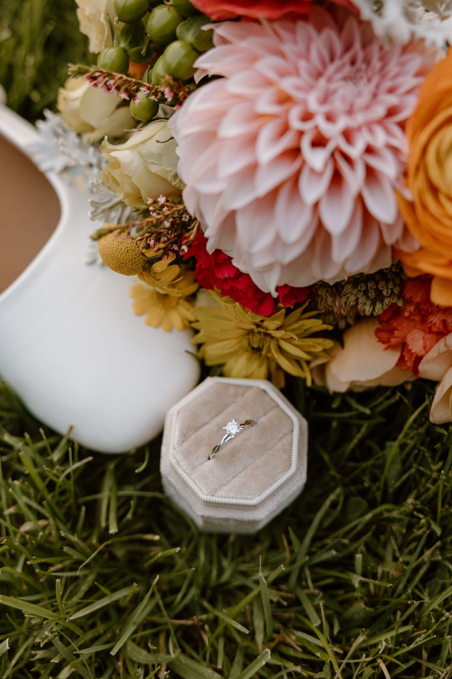 Wedding ring rests in a white velvet hexagonal box surrounded by pink, orange and green floral arrangements.