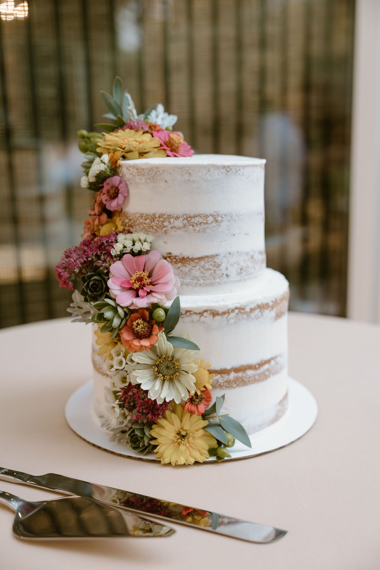 Two-tier semi-naked wedding cake decorated with cascading fresh flowers in pink, yellow and white on white cake stand.