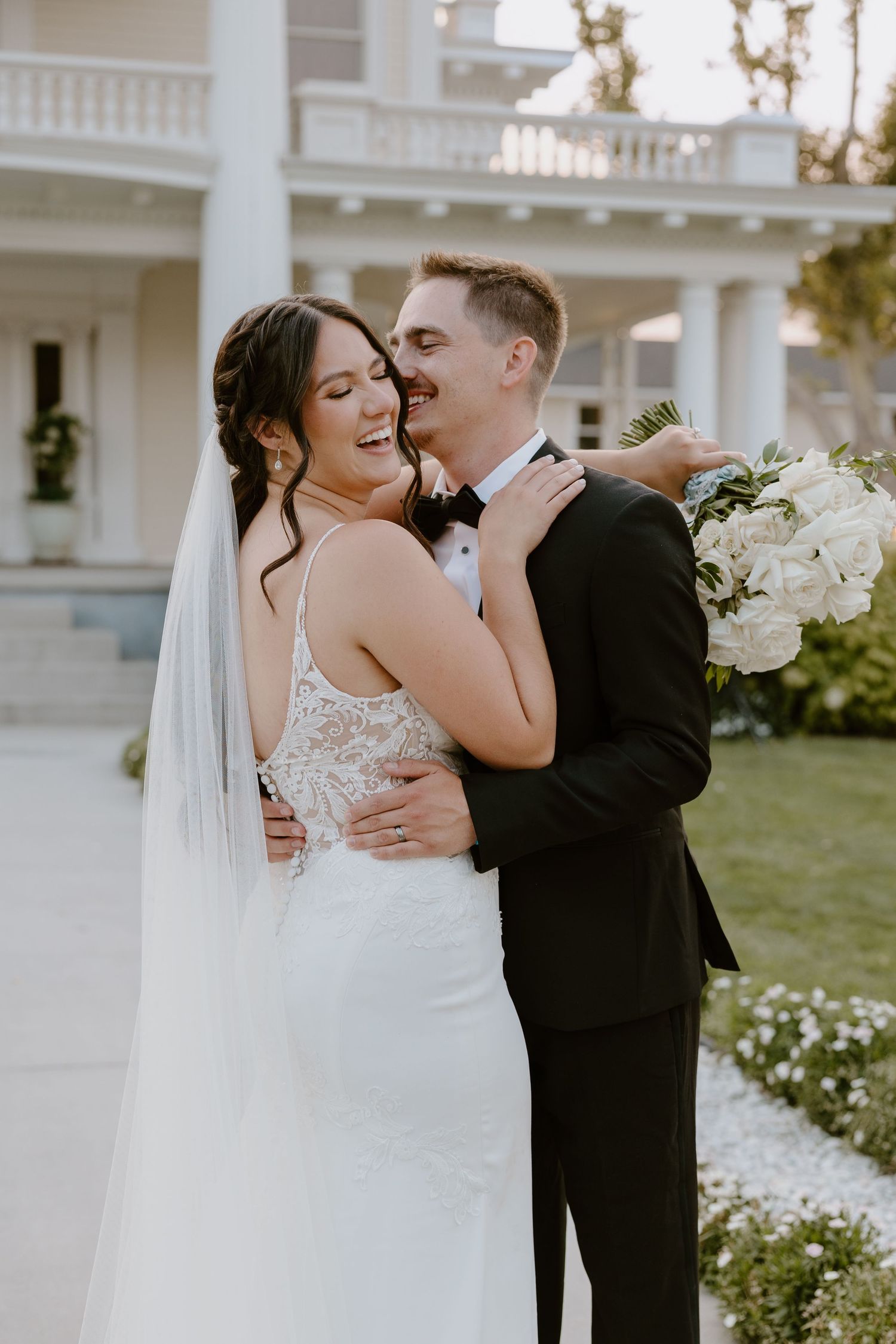 A romantic wedding couple shares tender moments on a sunny day in front of a white columned mansion.