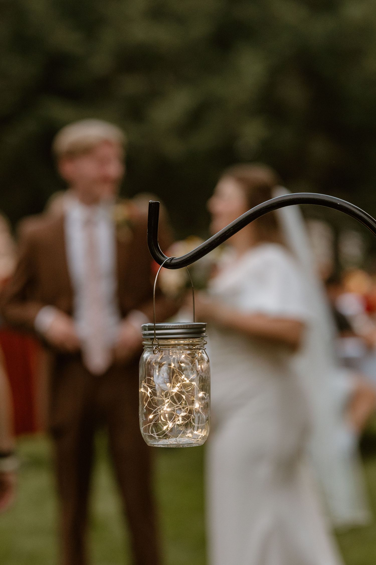 A glowing mason jar with fairy lights hangs from a tree branch at an outdoor wedding reception.