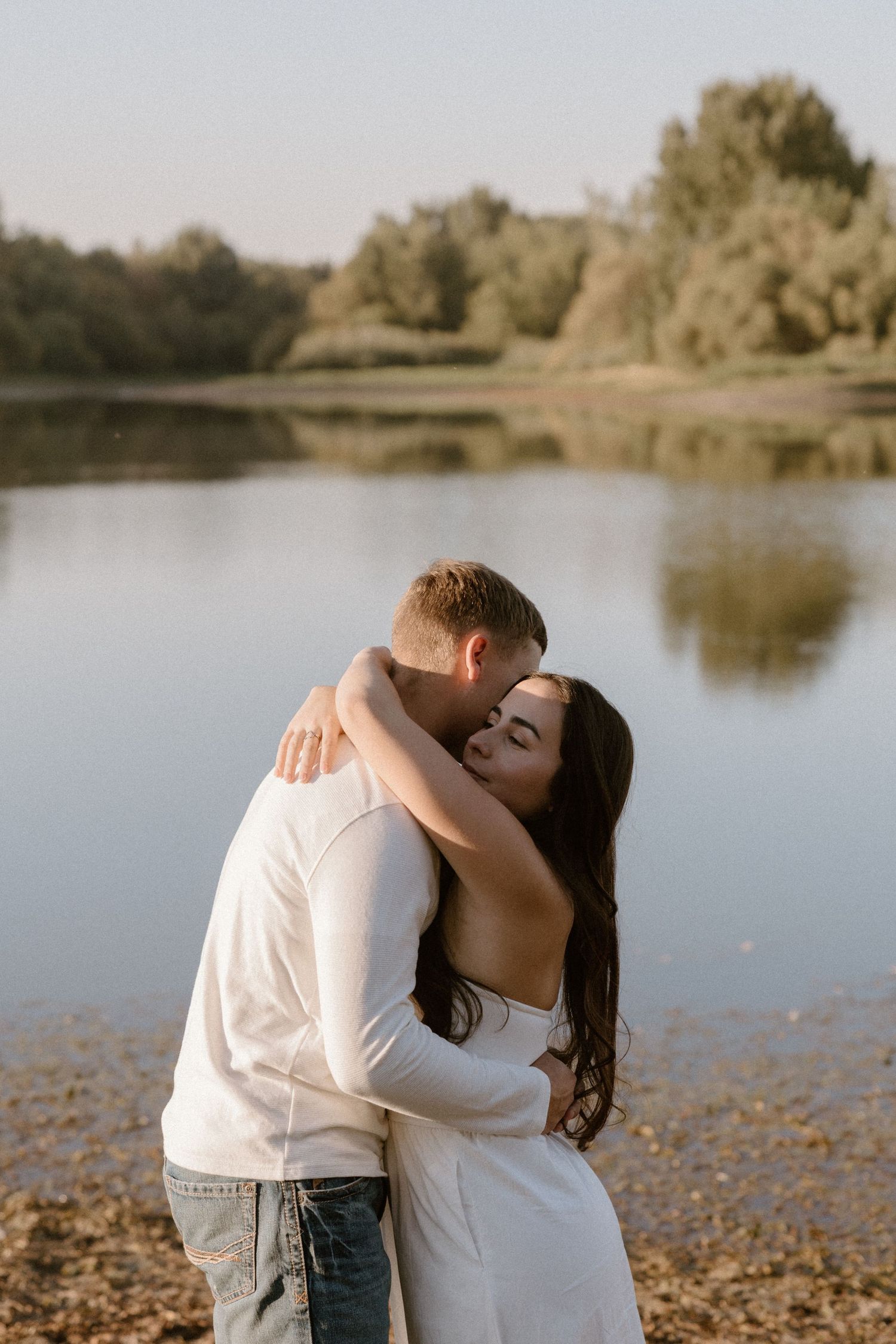 A romantic embrace by the tranquil lakeshore with trees reflecting in the calm water at sunset.