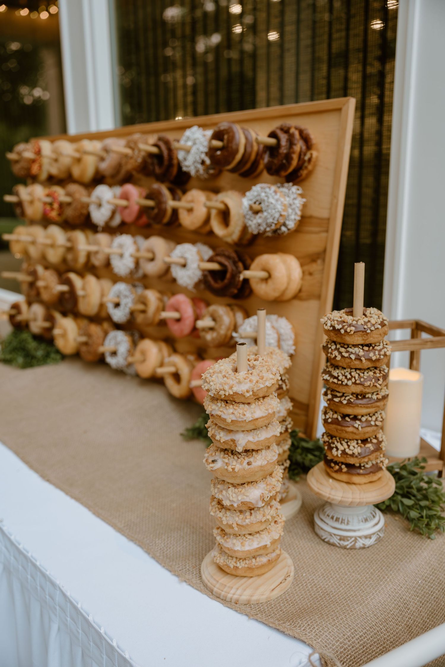 Stacked donuts with sprinkles and chocolate toppings displayed on wooden stands at a dessert table with burlap decor.