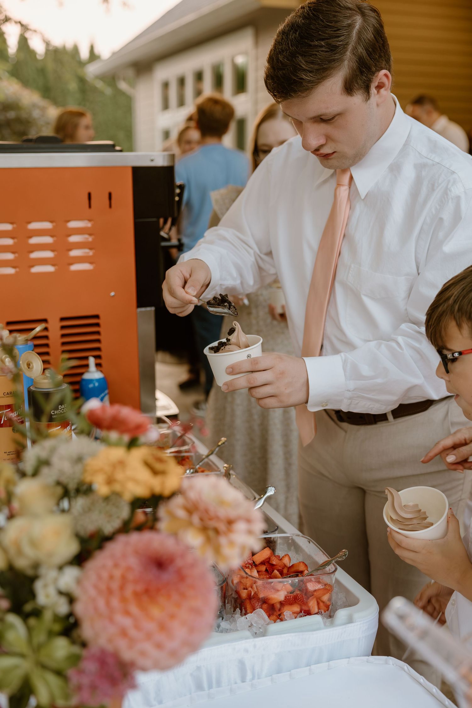 A person in a white shirt and peach tie serves ice cream into small bowls at an outdoor event.
