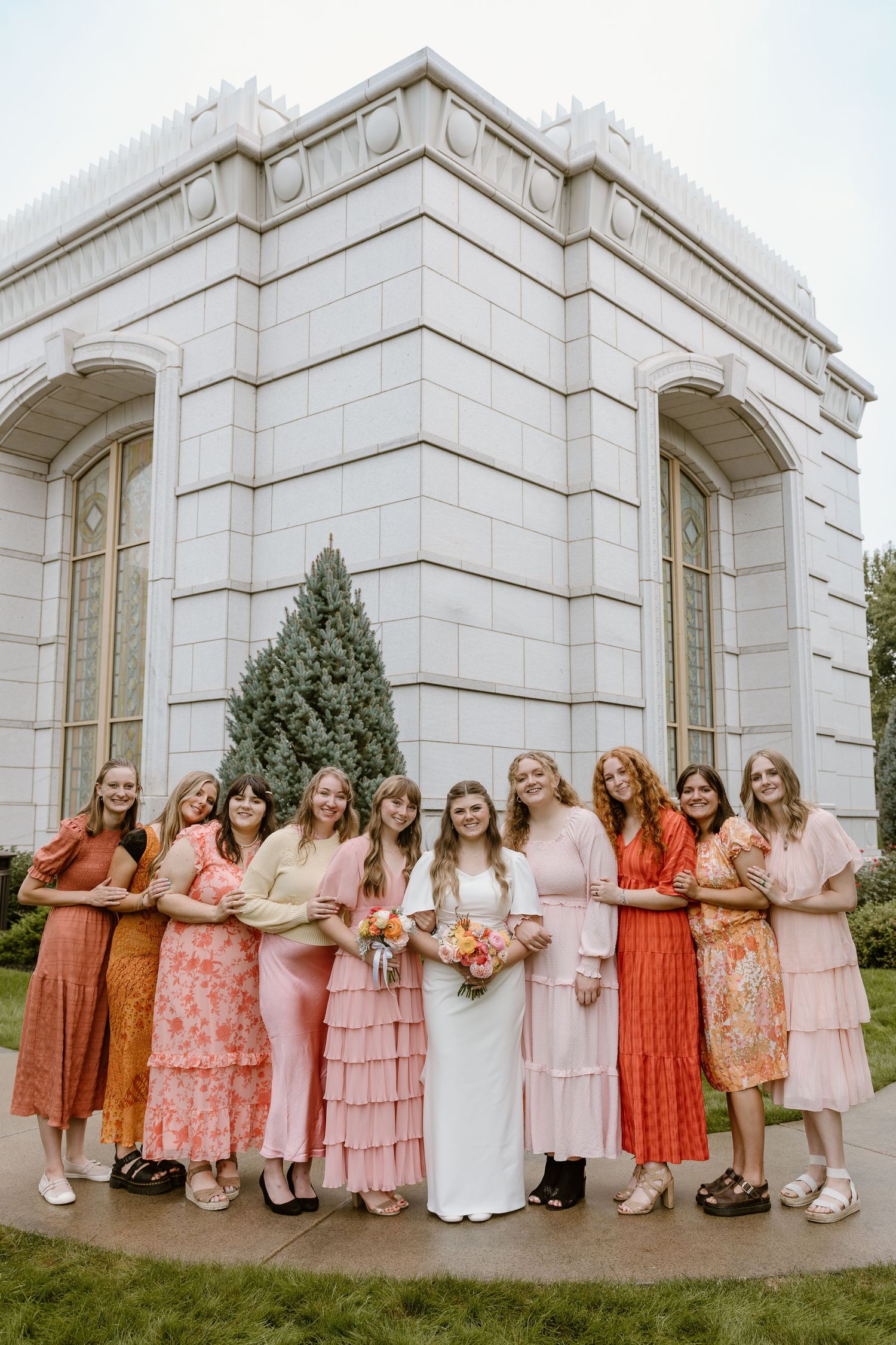 Group wearing coordinated pink and coral dresses pose together against white stone building exterior.