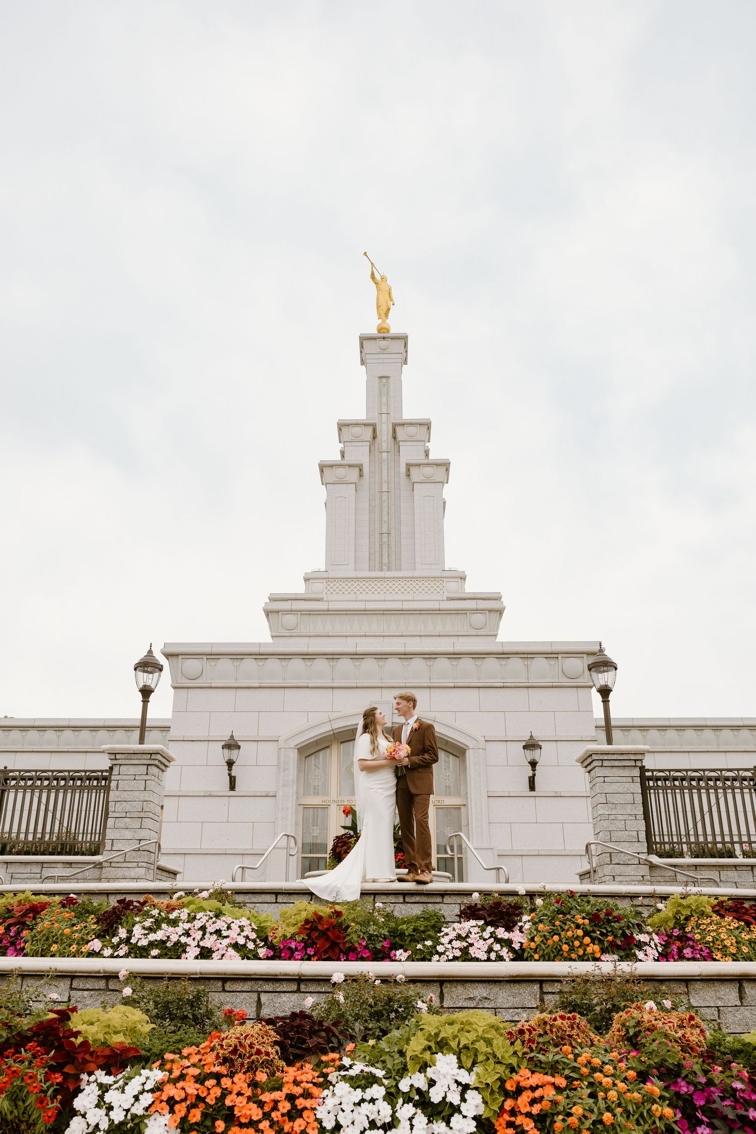 Colorful flower gardens with orange and white blooms surround a white Mormon temple building with ornate architecture.