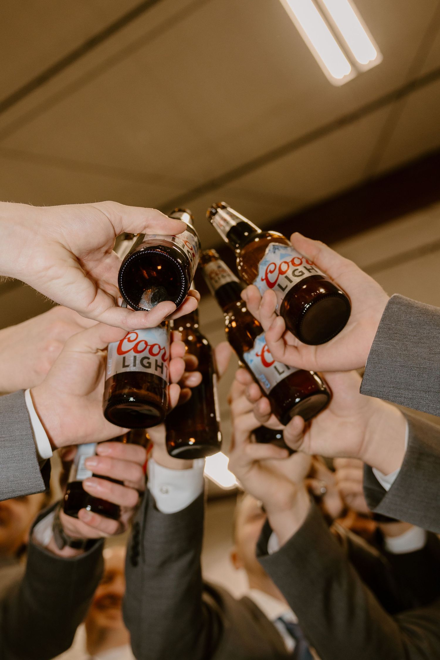 Group of hands toasting with Coors beer bottles in a warm-toned indoor setting with fluorescent lighting.