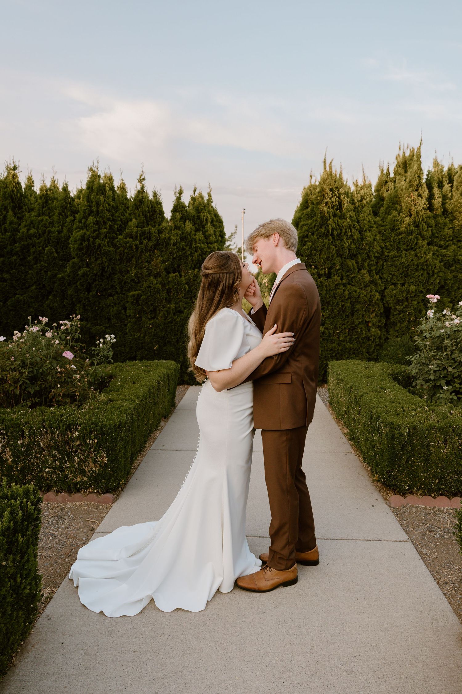 A romantic wedding couple embraces on a garden path lined with manicured hedges and flowers at sunset.
