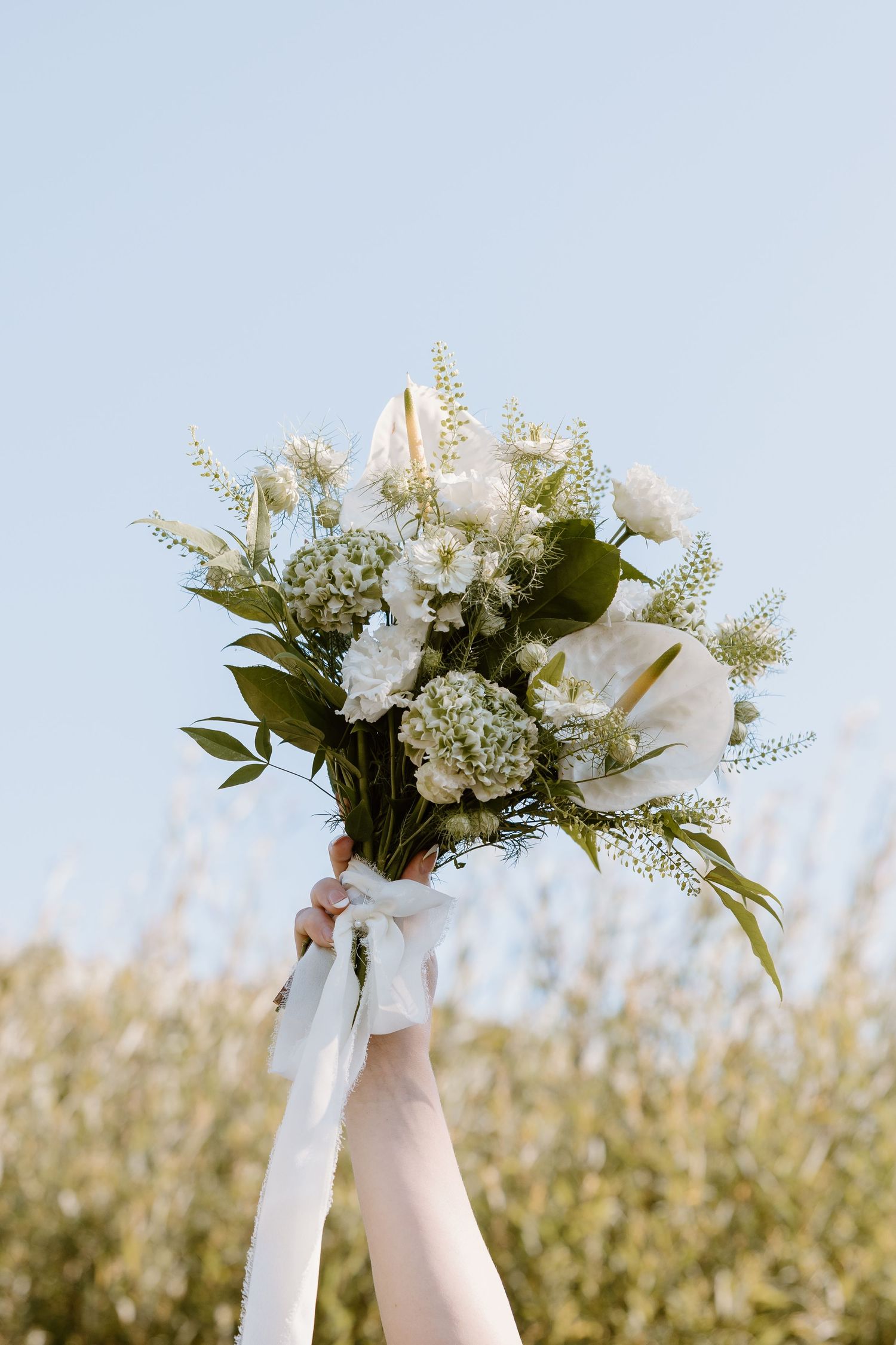 A white floral bouquet with lilies and delicate greenery held up against a soft sky background.