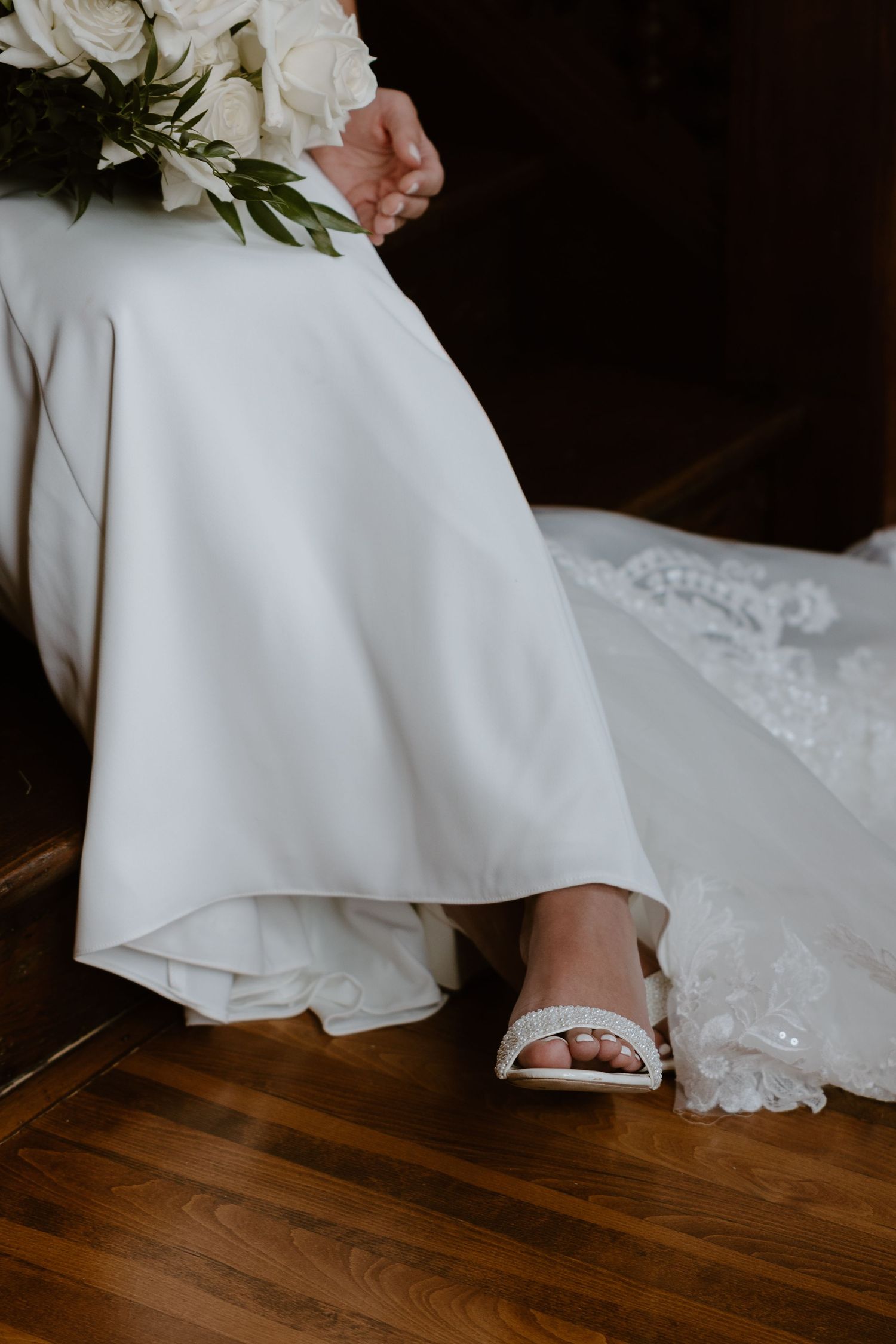Close-up view of a white wedding dress hem and delicate lace details against a dark hardwood floor.