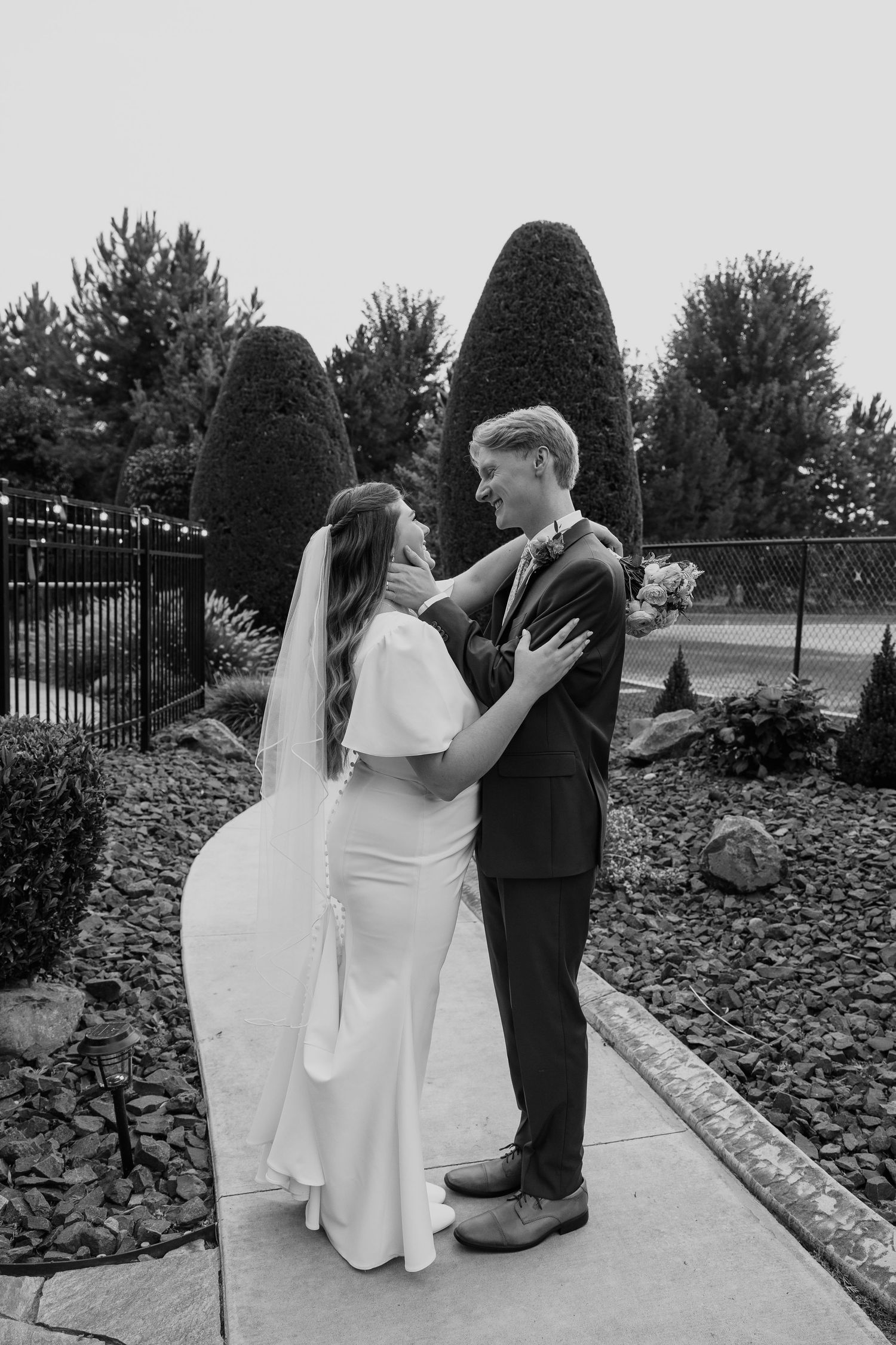 A couple in wedding attire embraces on a garden path with sculpted shrubs and a decorative fence in the background.